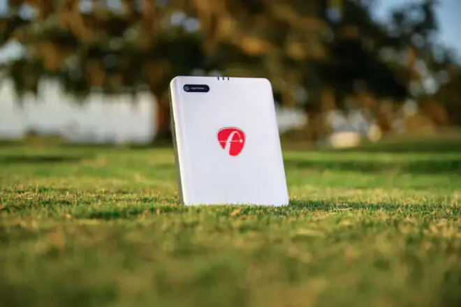 A white golf ball retriever with a red logo stands upright on a grassy field with trees in the background.
