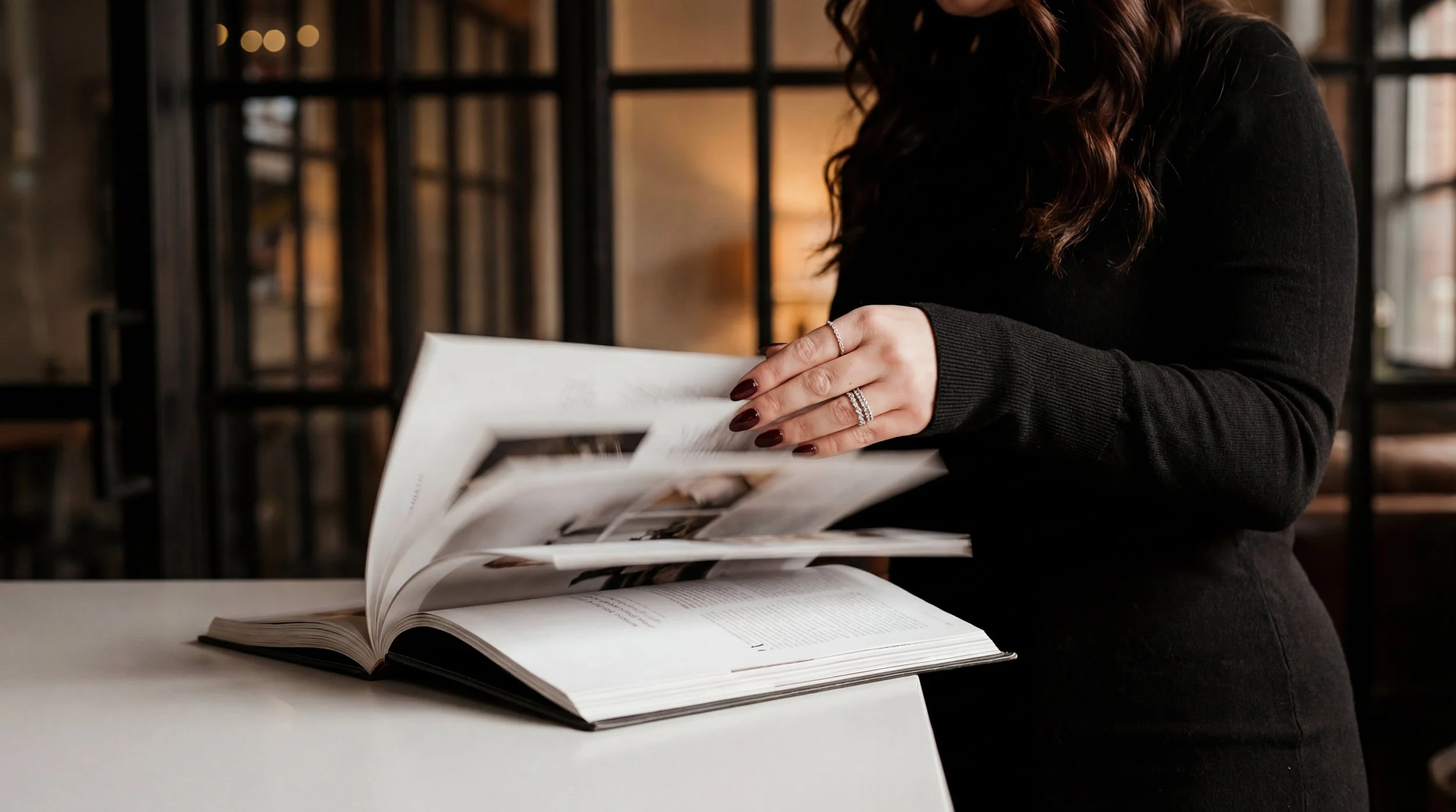 A woman with dark curly hair, wearing a black long sleeve top and rings, turning pages of a large open book on a white table in a modern, industrial-style room.