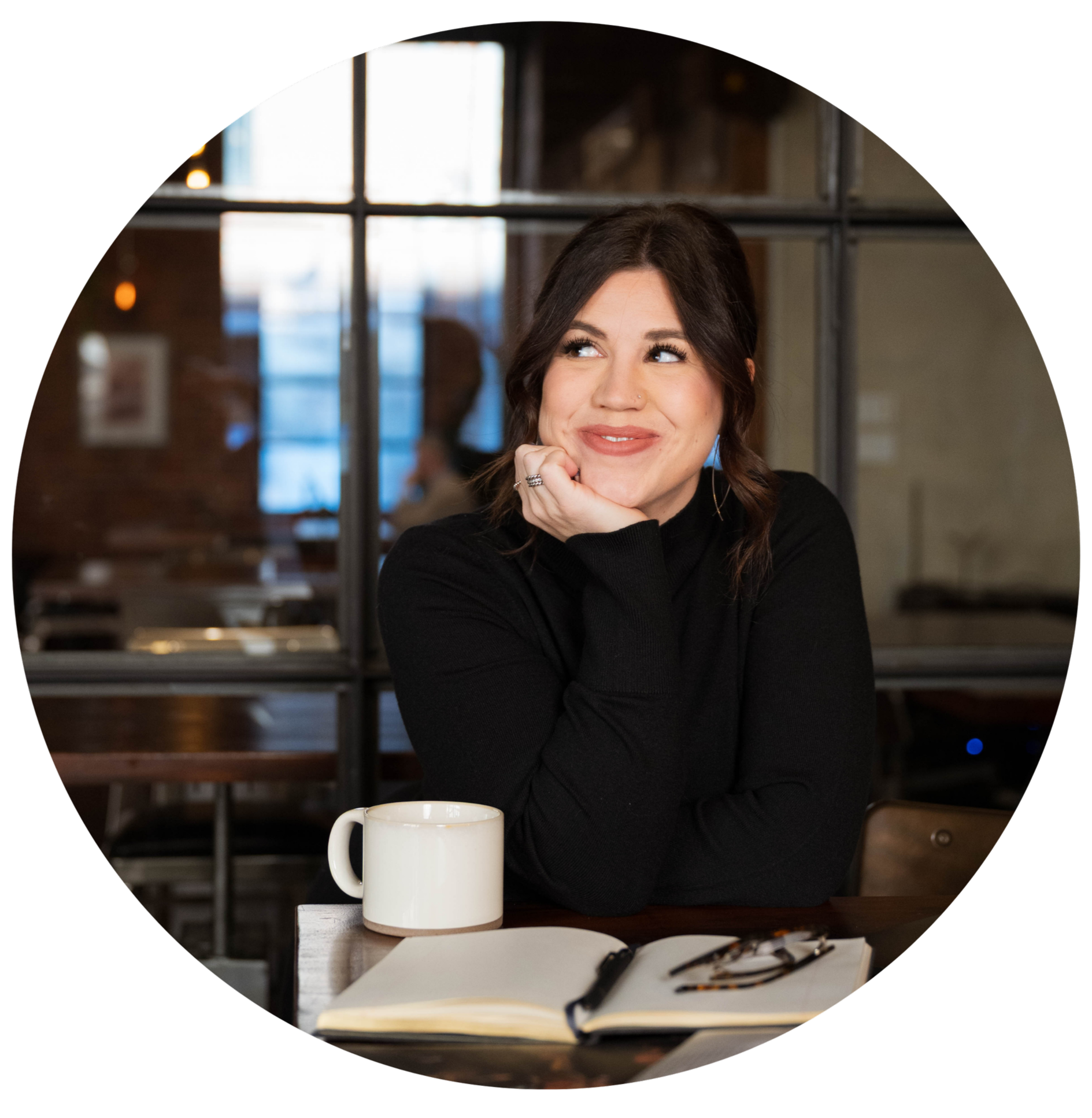 A woman with dark hair, wearing a black top, sits at a table with her chin resting on her hand, smiling and looking to the side. A white mug, open book, and glasses are on the table. The background features a window with black framing, and warm indoor lighting.