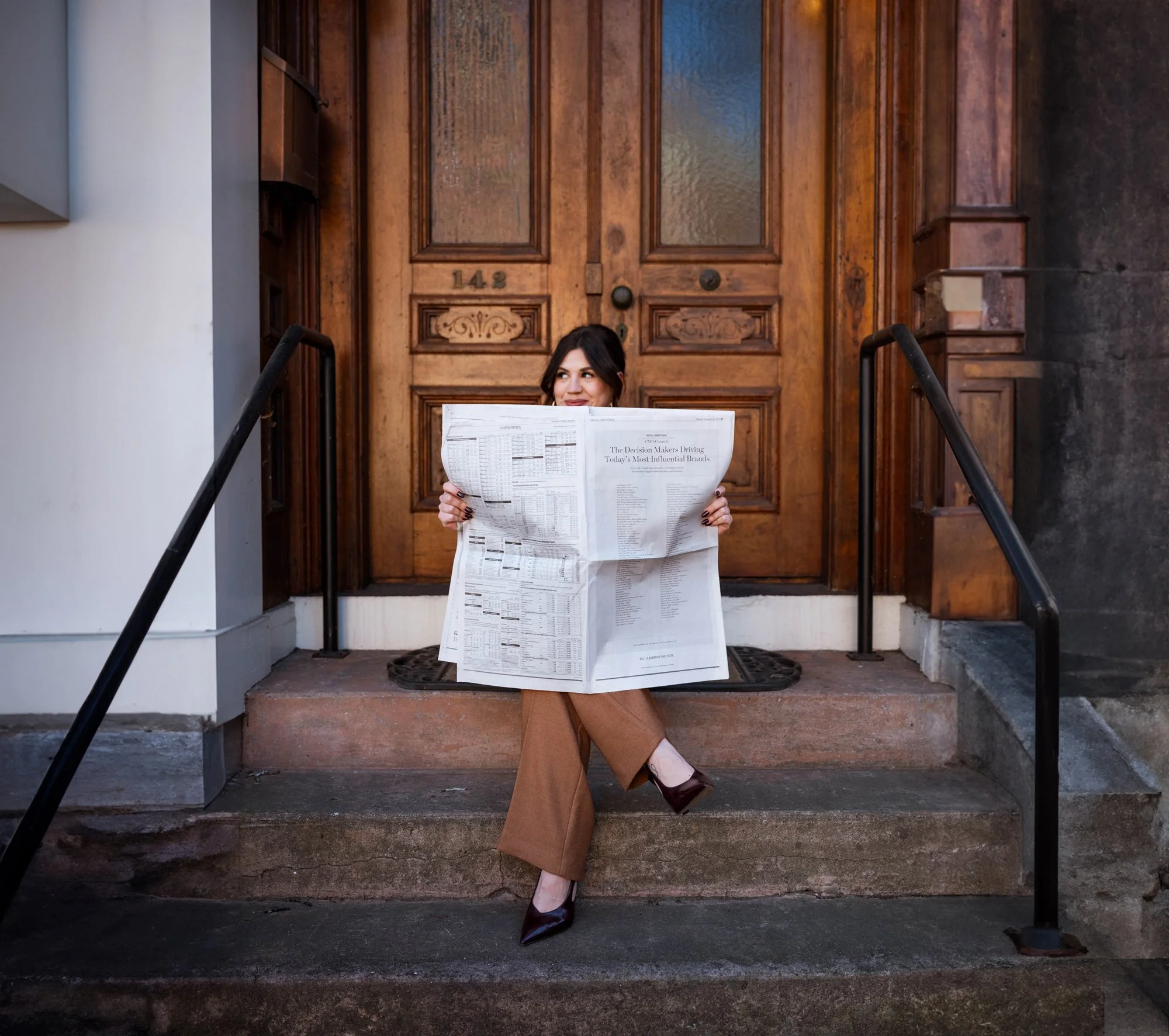 Woman sitting on steps in front of a wooden door, reading a large newspaper, wearing brown pants and heels.