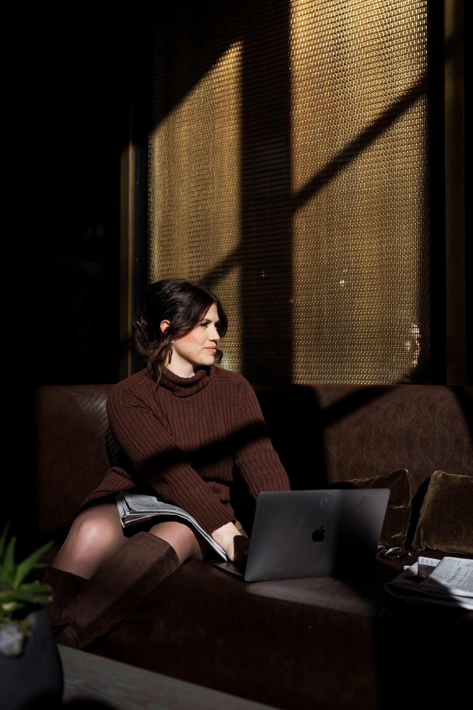 Female Therapist taking notes while reflecting on practice alignment, sitting on a brown sofa with a laptop and newspapers in a dimly lit room, illuminated by sunlight coming through a textured, metallic grid pattern window.