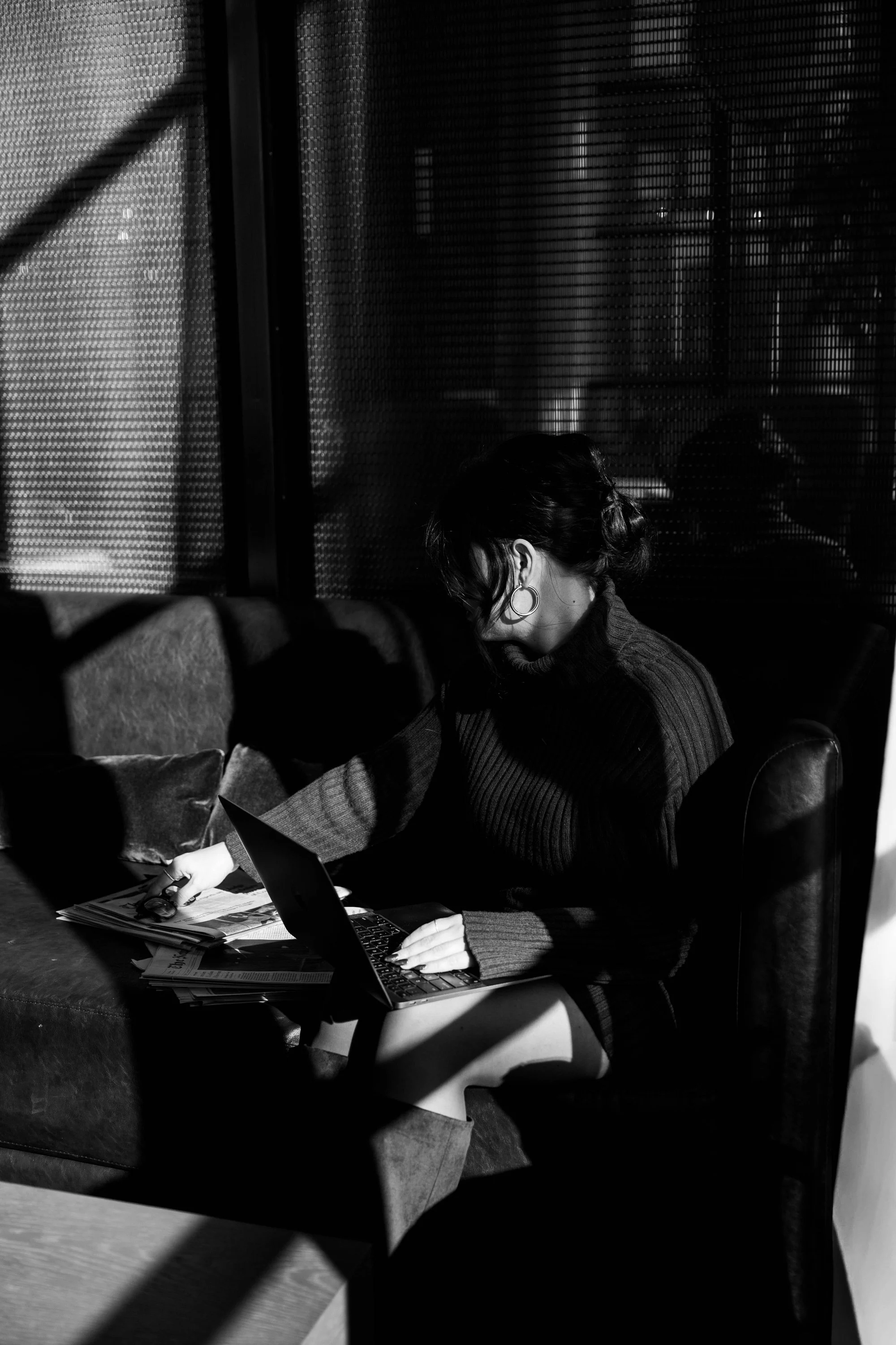 A woman sitting on a couch, working on a laptop, with newspapers and magazines around her, in a dimly lit room with curtains and textured walls.