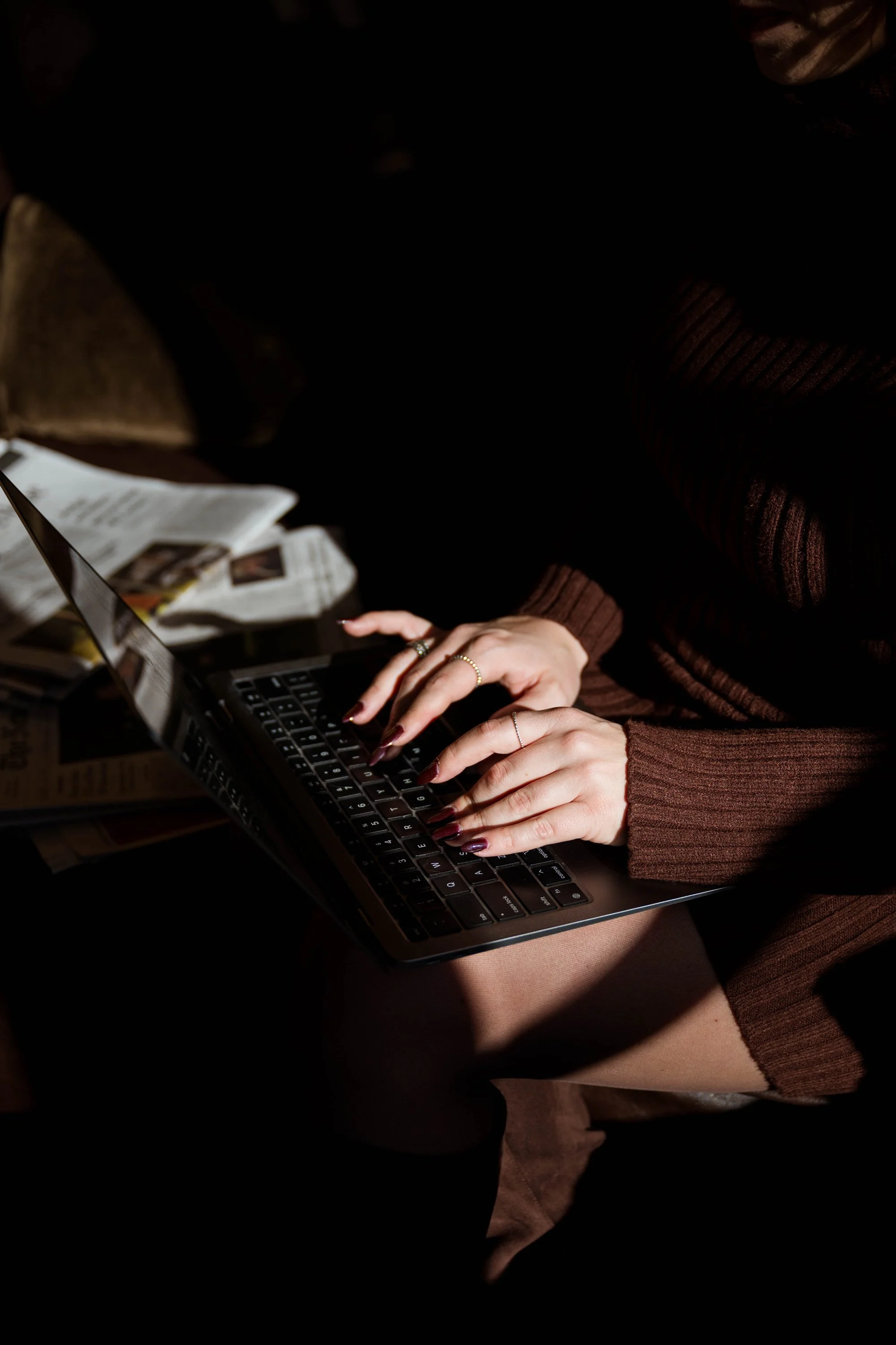 Person typing on a laptop in a dimly lit room, wearing a brown sweater.