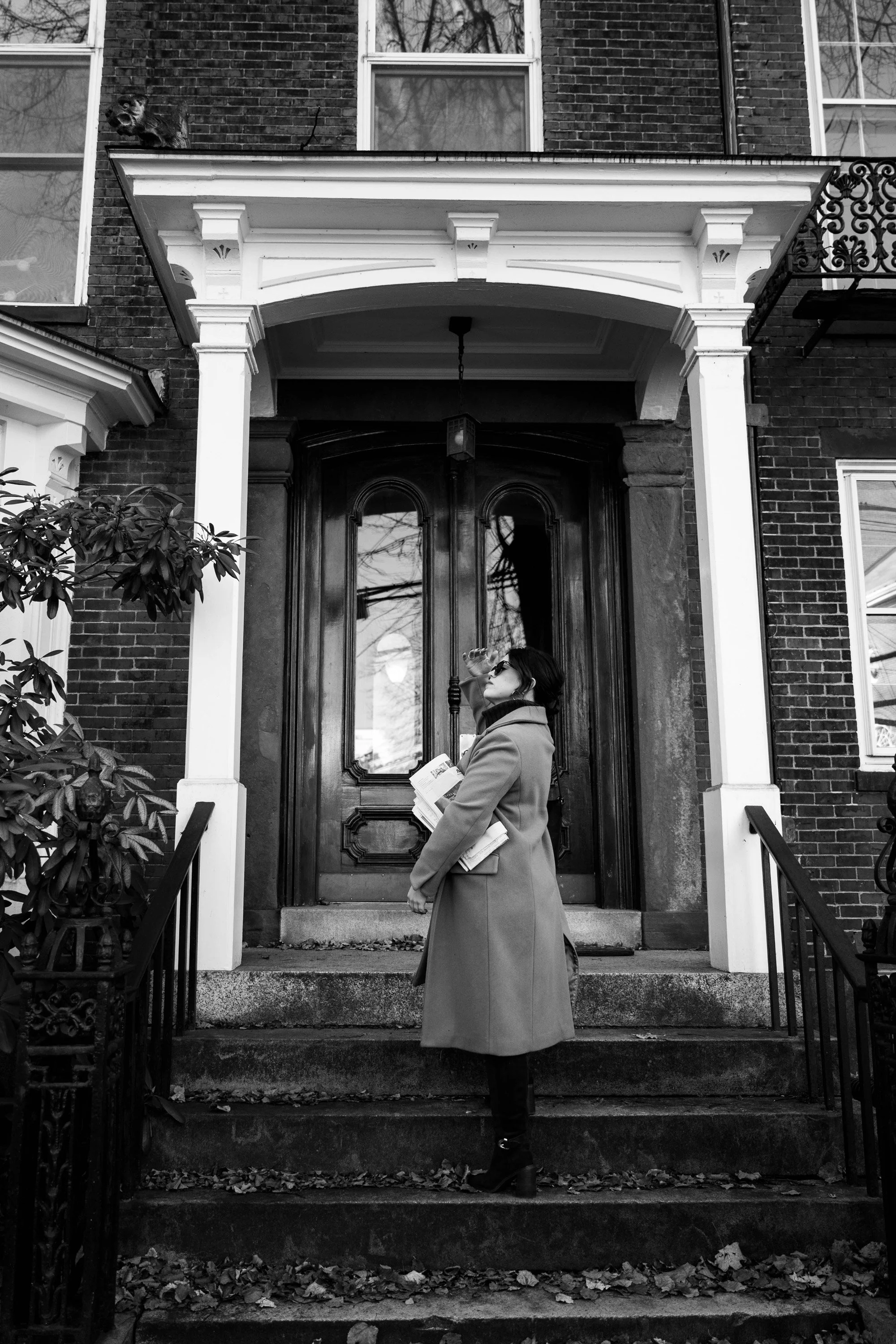 A woman in a coat and sunglasses standing on front steps of a house, drinking water from a bottle and holding newspapers.