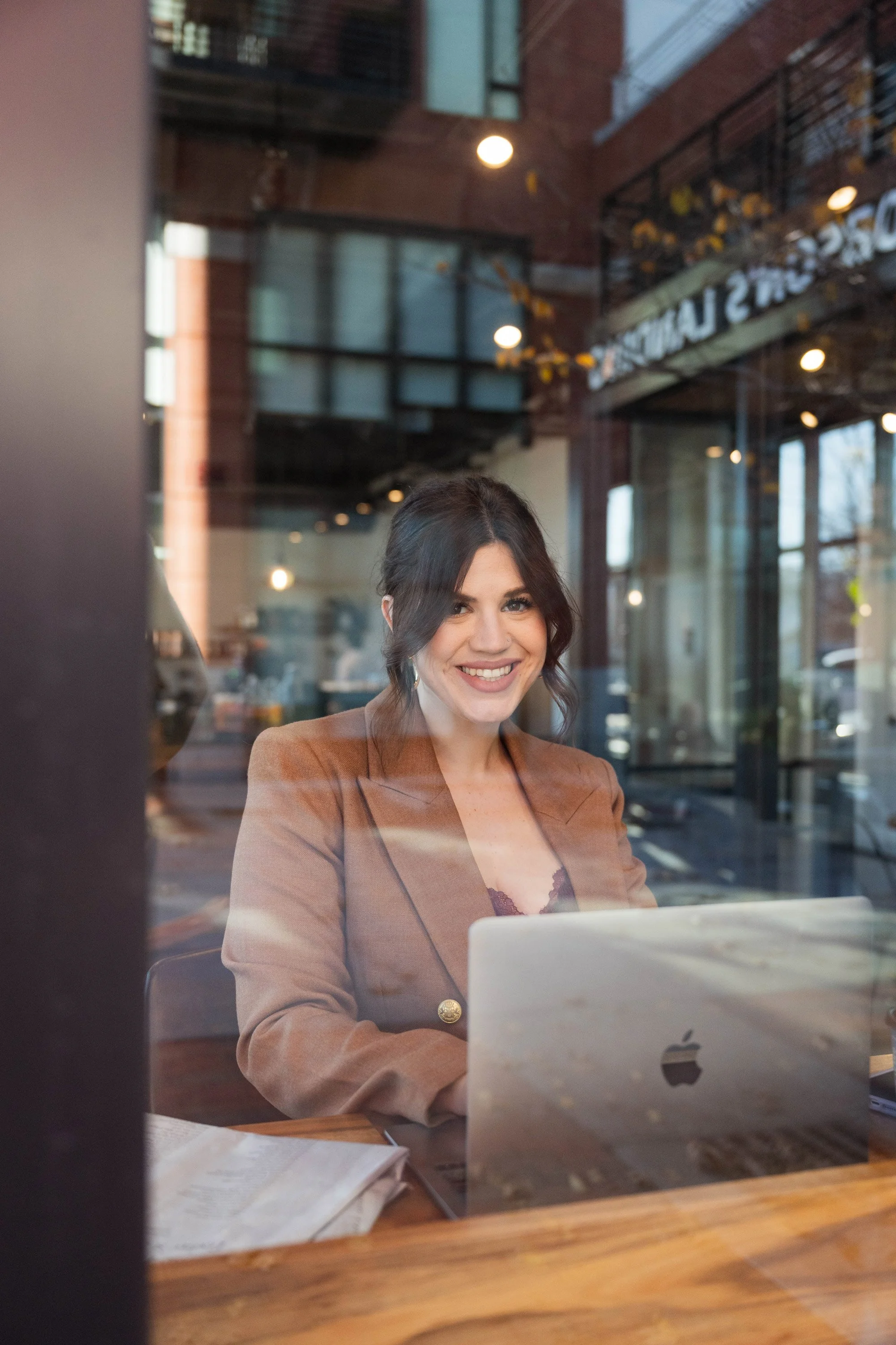 A woman smiling while working on a laptop inside a cafe, viewed through a window.