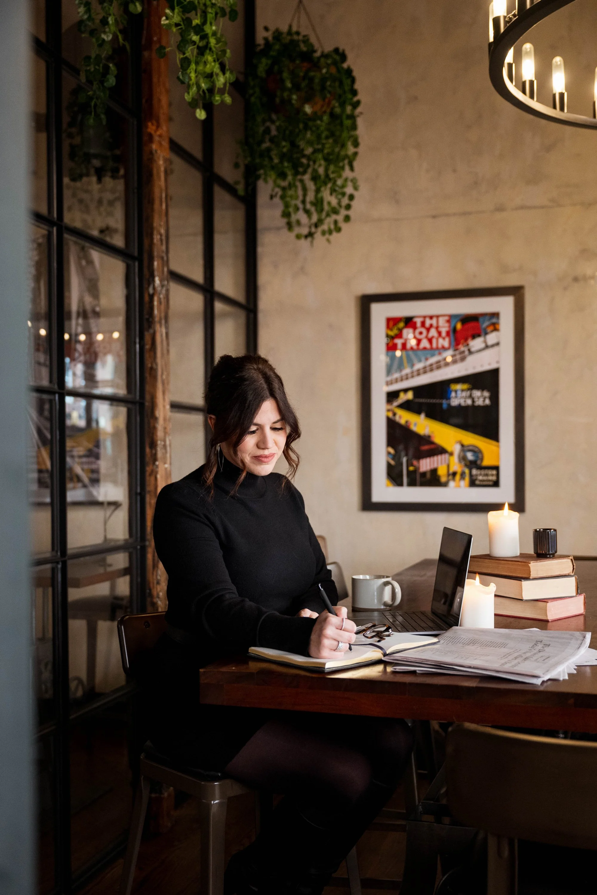 A woman sitting at a wooden table in a cozy, warmly lit room, writing in a notebook with a laptop, candles, and stack of books on the table, with framed artwork on the wall behind her.