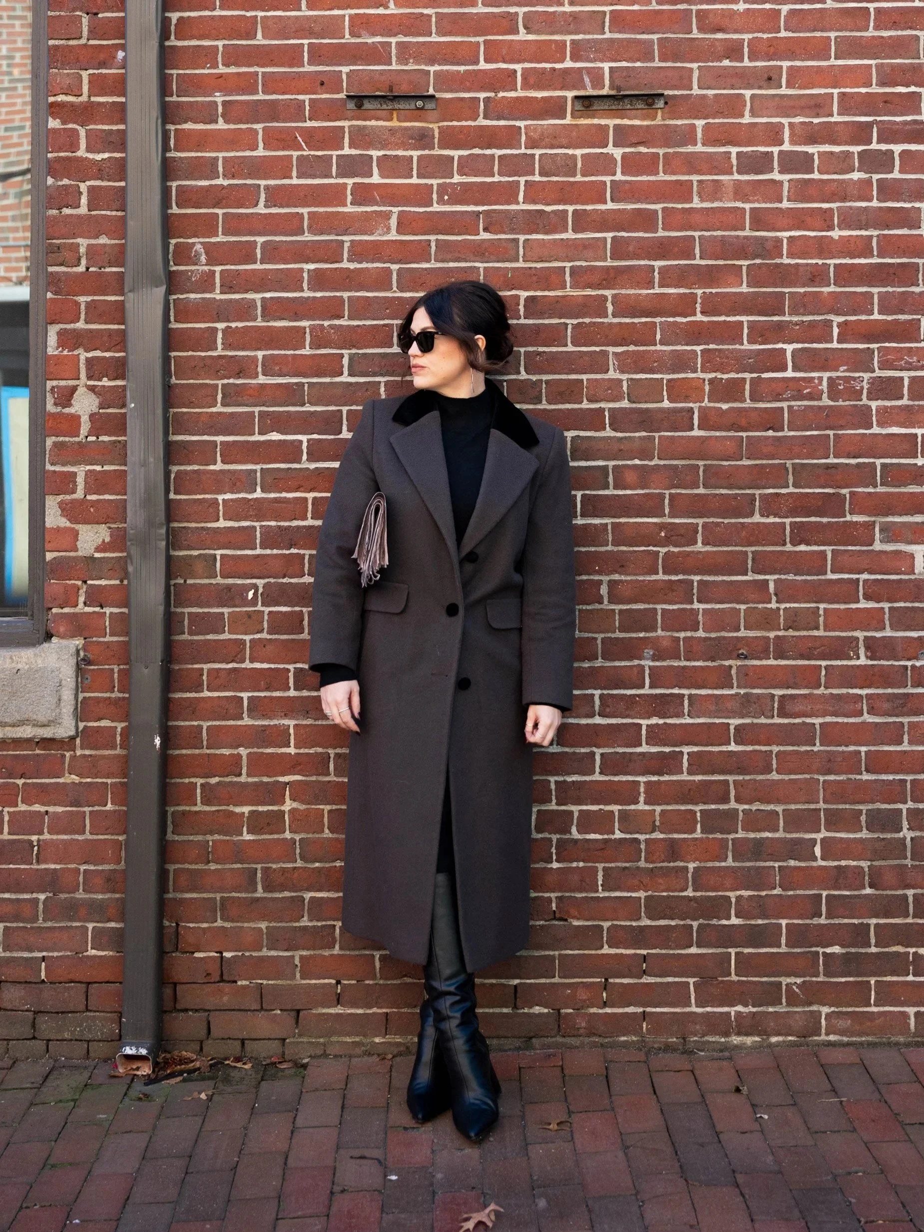 Anna Olsen, LMHC coach and founder of The Fulfilled Therapist standing against a brick wall, wearing a long gray coat with black collar, black turtleneck, black pants, black knee-high boots, sunglasses, and carrying a gray handbag with fringe.
