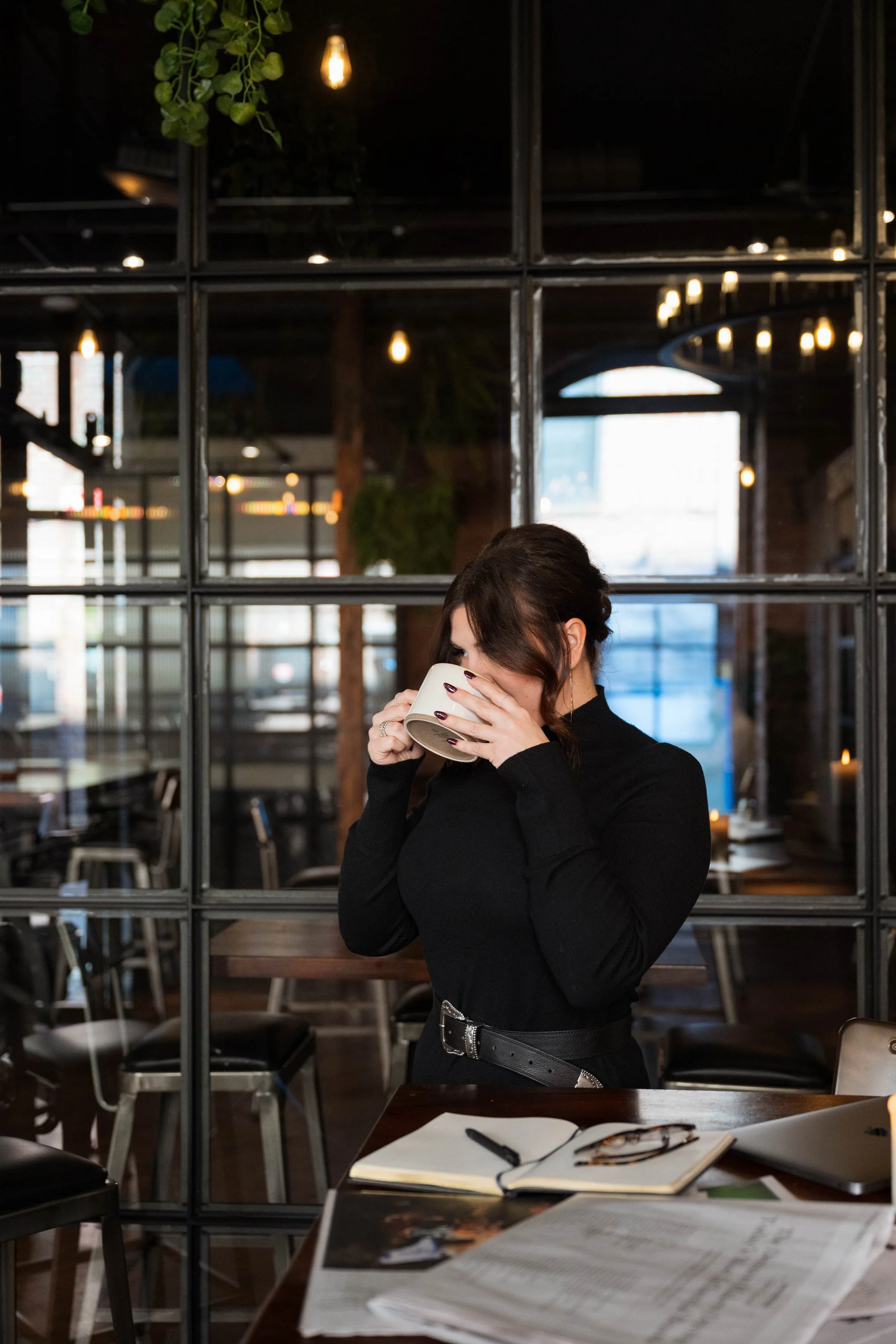 A woman in black clothing drinking from a white mug inside a cozy, modern restaurant or cafe with warm lighting, wooden furniture, and industrial-style decor.