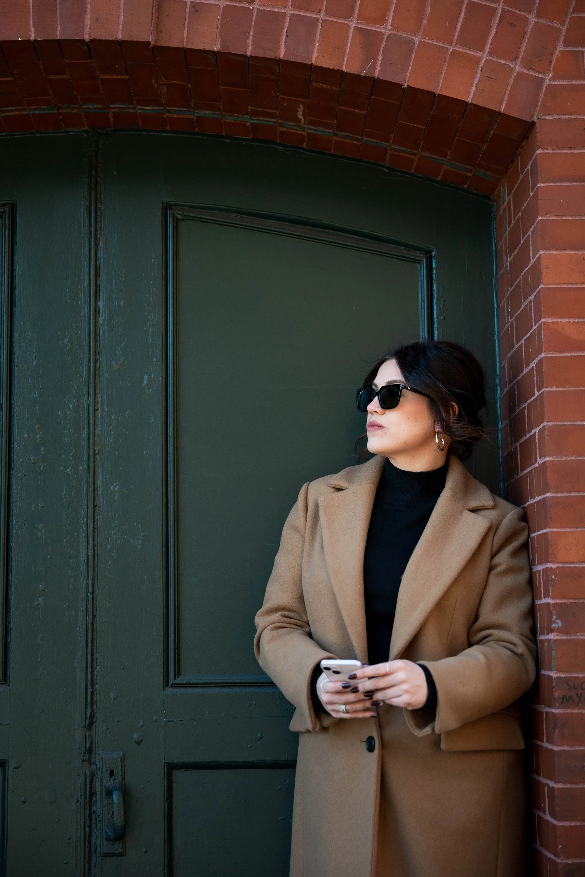 Anna Olsen with dark hair wearing sunglasses, a tan coat, and a black turtleneck, holding a smartphone, standing outside near a green door and brick wall.