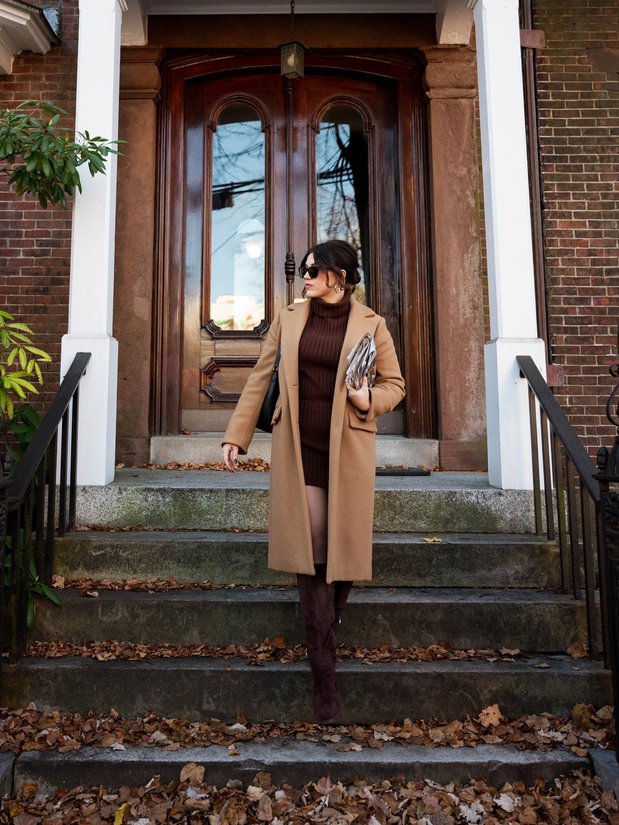 Woman in brown coat and sunglasses walking down front steps of a brick house, holding papers, autumn leaves on the ground.