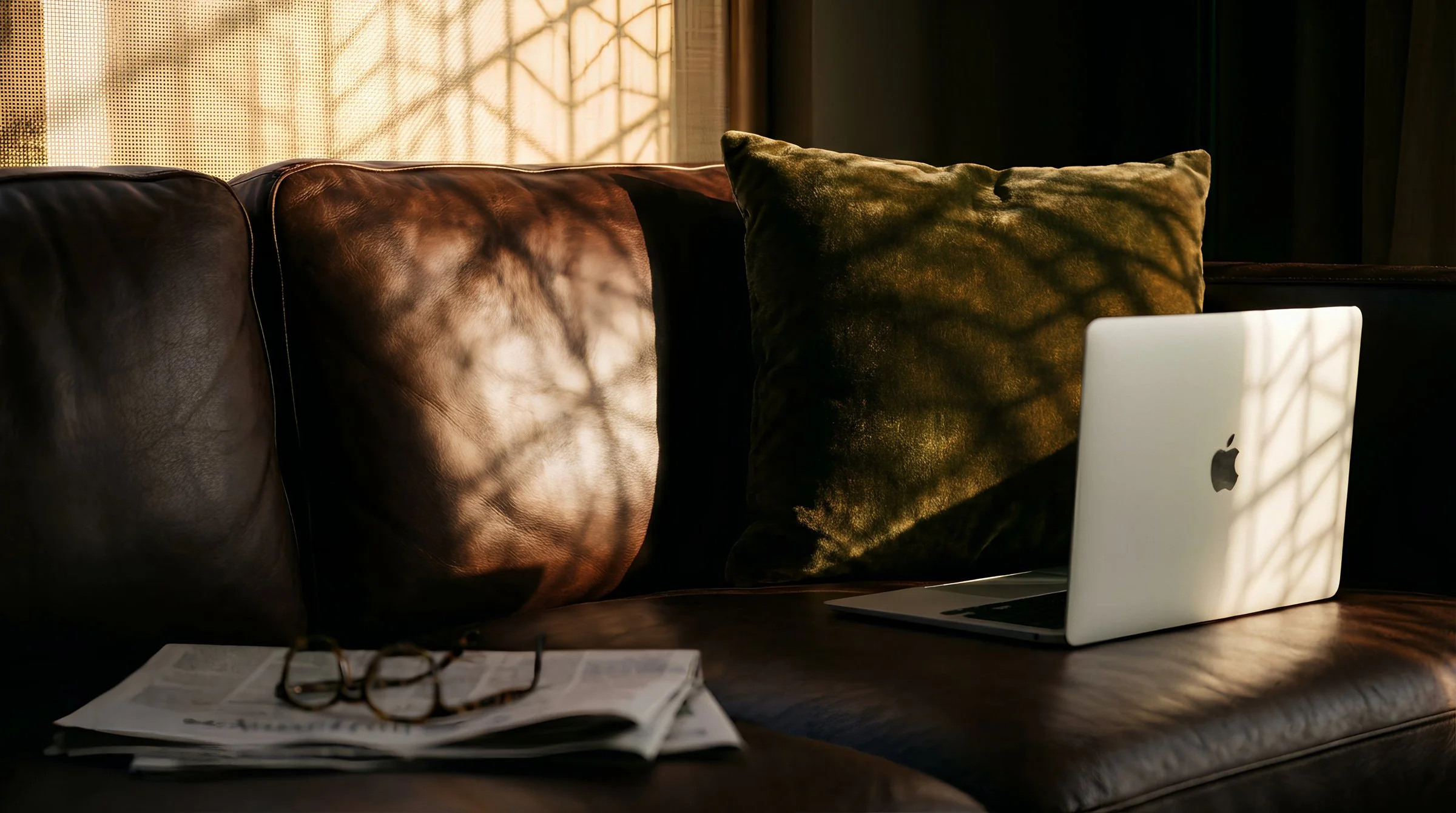 A leather sofa with a green pillow, a pair of glasses, and an open laptop on top, with sunlight creating a shadow pattern on the pillow and laptop.