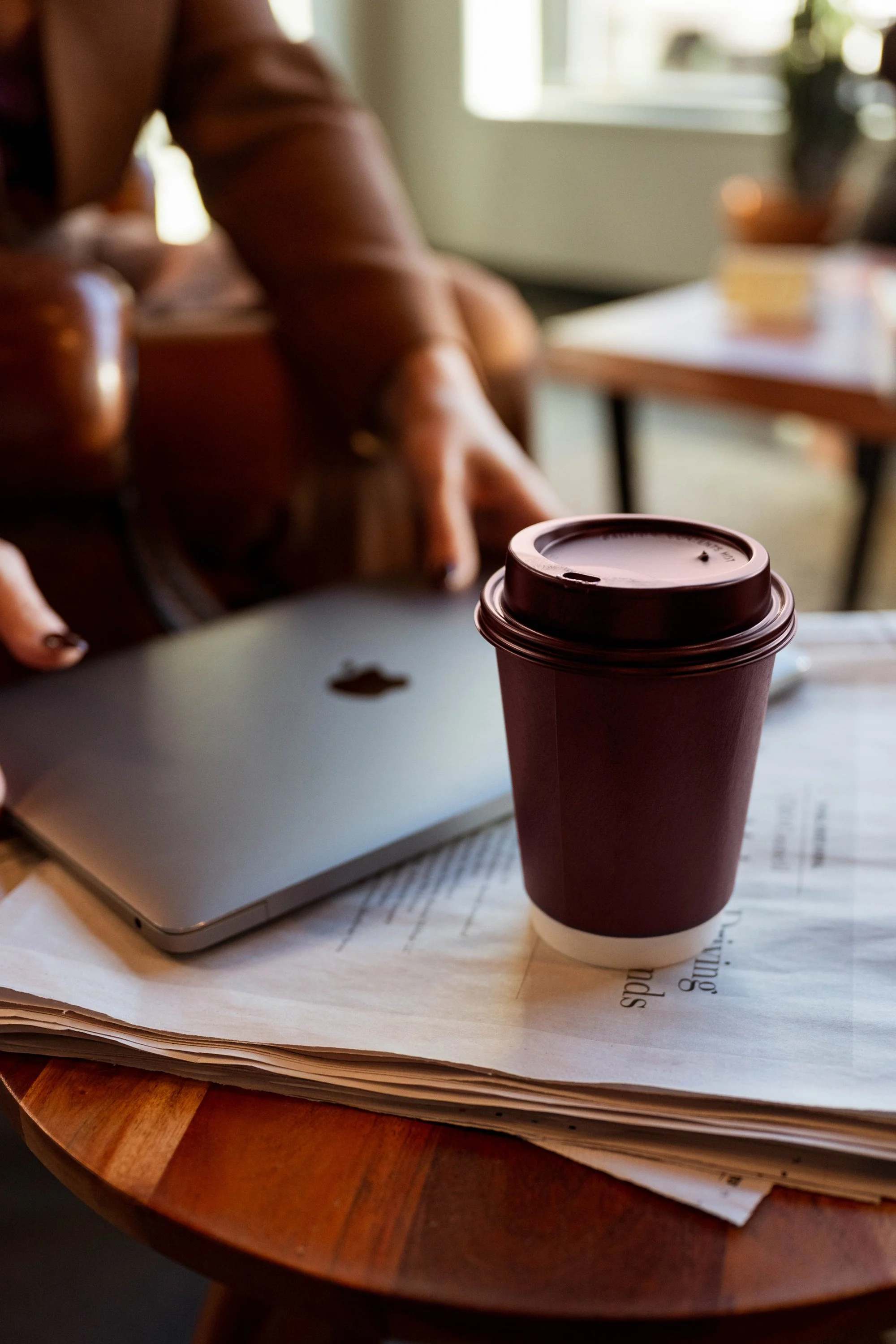 A to-go coffee cup on top of a stack of papers on a wooden table, with a closed laptop and a person sitting in the background.