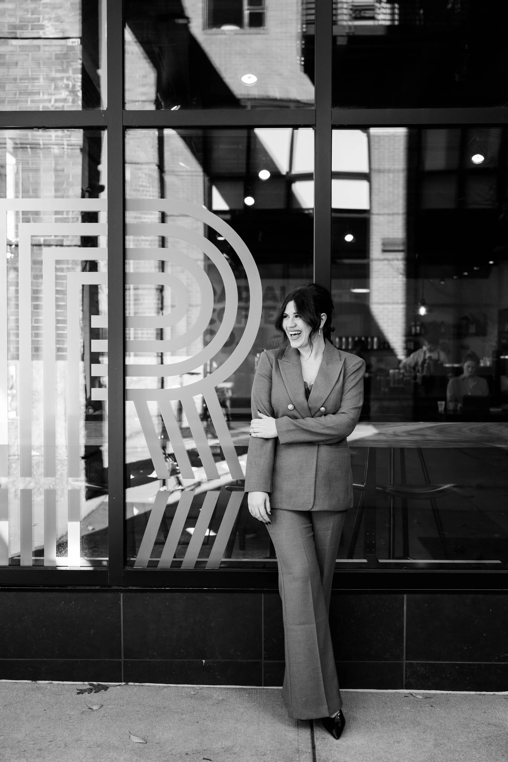A woman in a business suit laughing outside a modern cafe or restaurant with large glass windows and an abstract design on the window. The scene is in black and white.