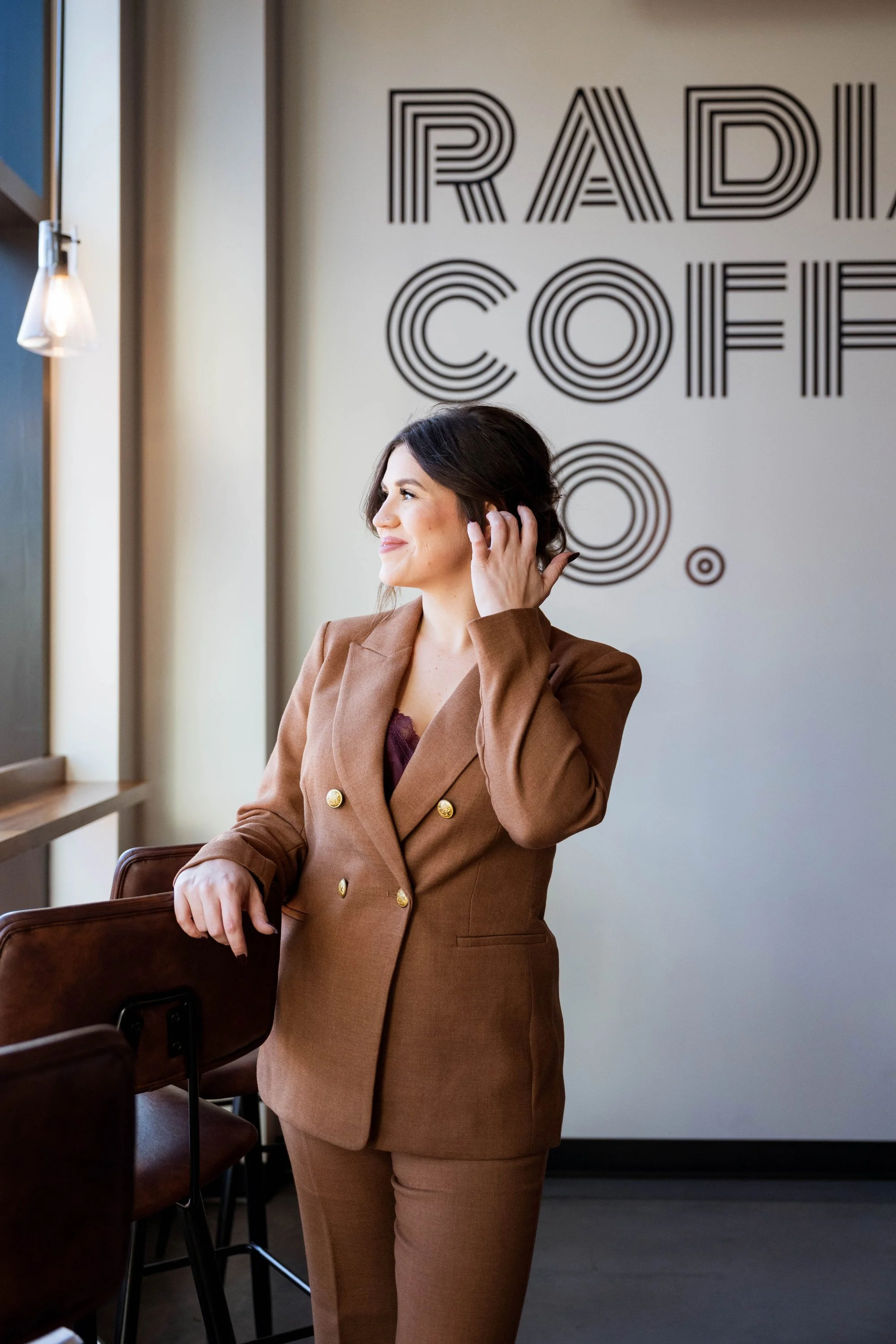 A woman, Anna Olsen, LMHC, in a brown suit standing inside a coffee shop called Radio Coffee Co., smiling while touching her ear and looking out the window.