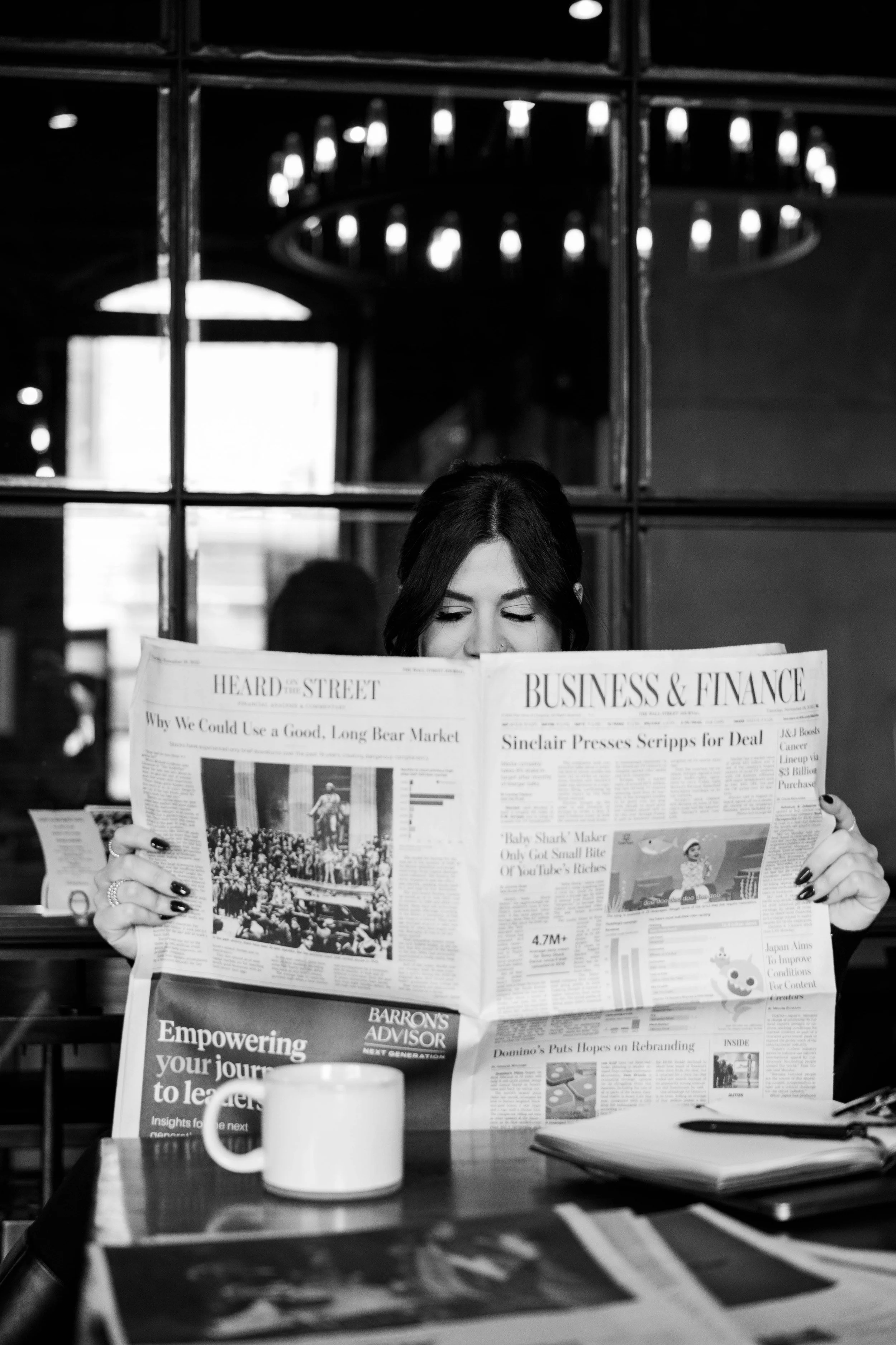 A woman reading a newspaper at a table in a dimly lit room with a chandelier overhead.