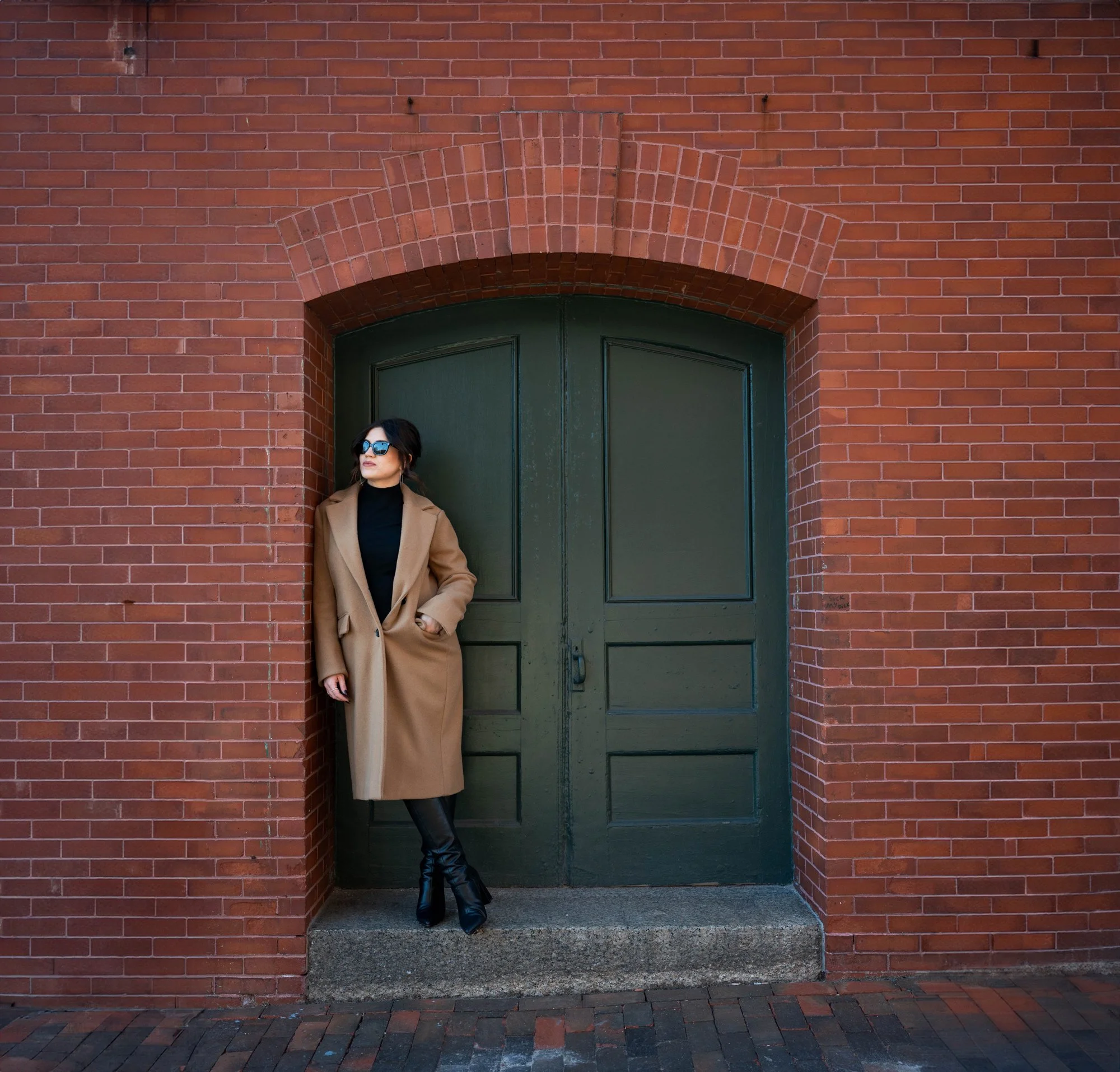 Anna Olsen, therapist, coach and founder of The Fulfilled Therapist, Woman in a tan coat and black boots leaning against a brick wall with a large green door.