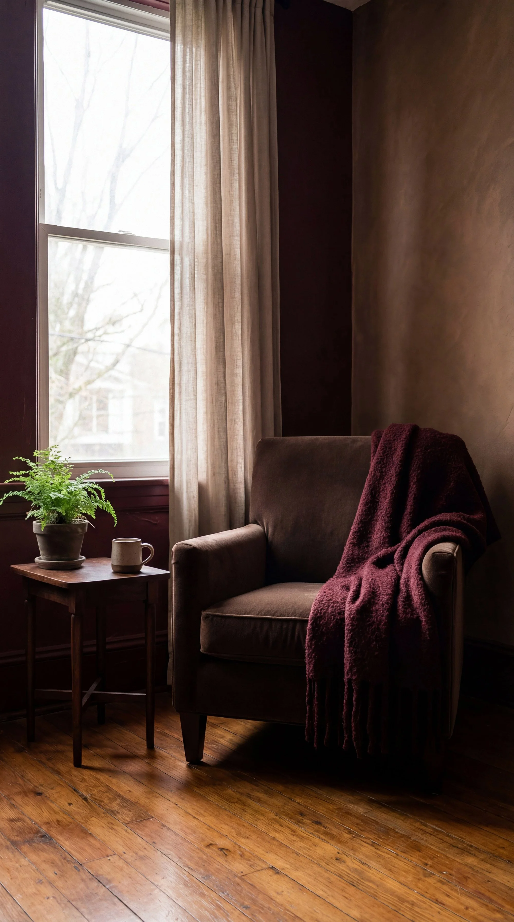 A cozy corner with a brown armchair draped with a burgundy blanket, next to a small wooden side table holding a green potted fern and a beige mug. There's a large window with sheer curtains allowing natural light into the room, and the floor is made of wood.