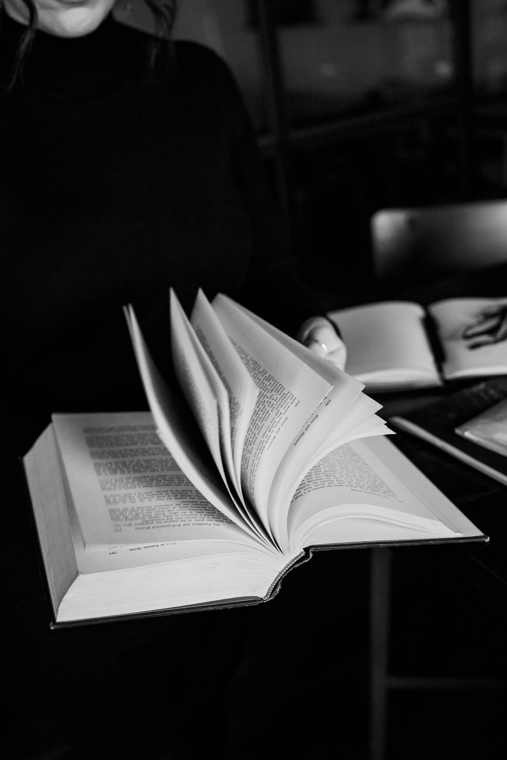 Open book with pages turning, on a dark desk, with another open book and a laptop nearby, in a dimly lit setting.