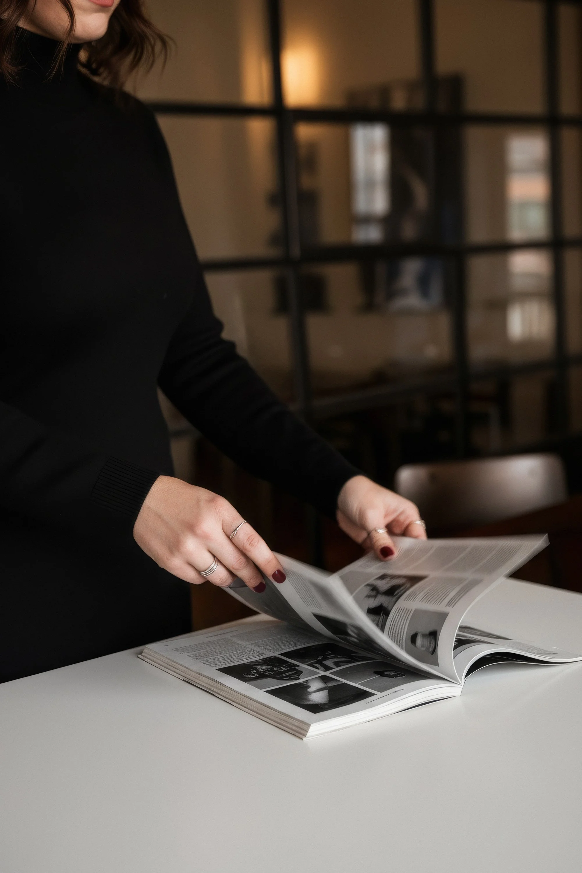 A woman with dark hair, dressed in a black long-sleeve top, is flipping through a magazine on a white table in a modern, dimly-lit room with a large window or glass wall behind her.