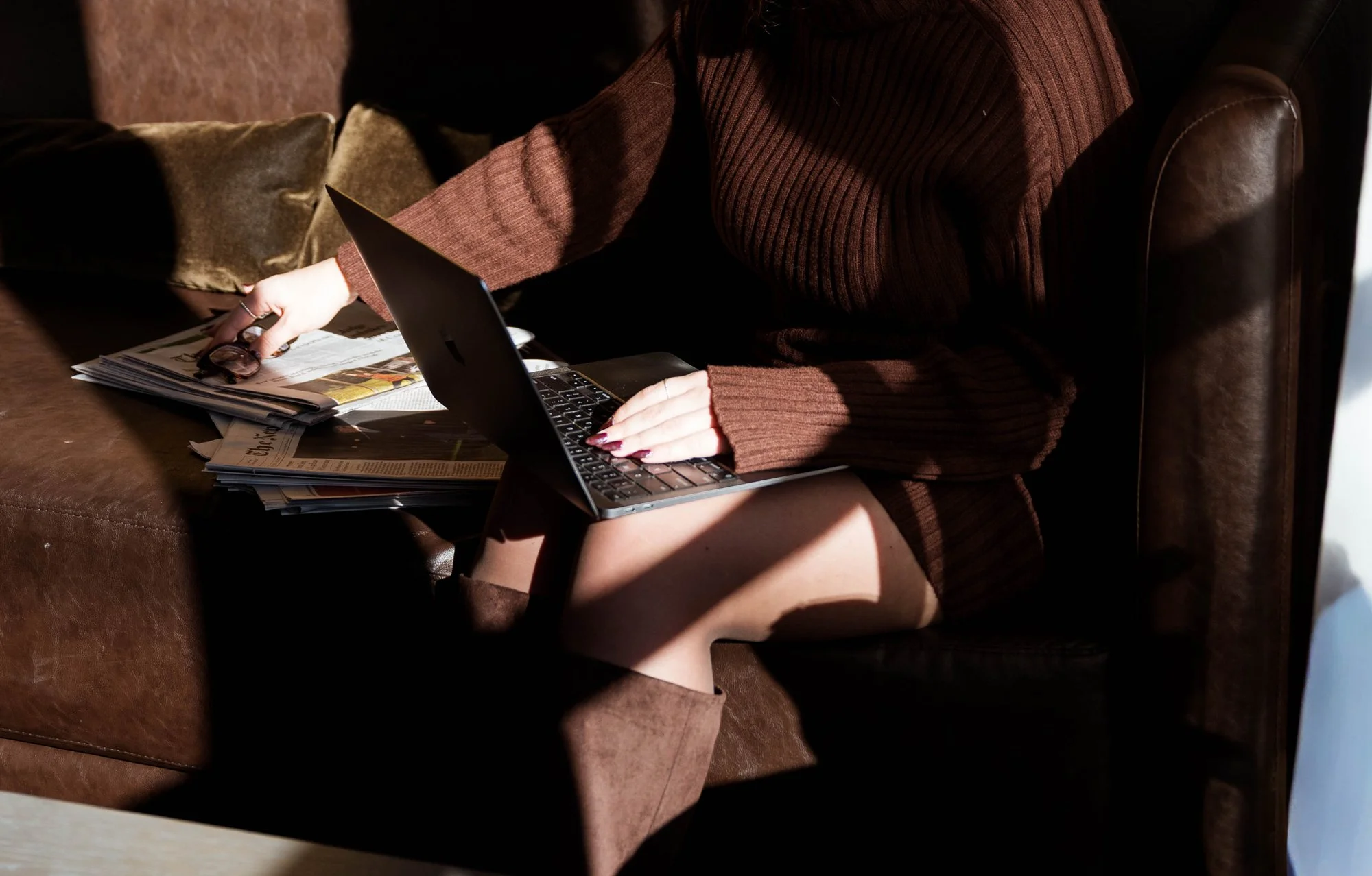 A person sitting on a brown leather sofa, wearing a brown sweater and boots, holding a laptop on their lap with a stack of newspapers and a pair of glasses nearby.