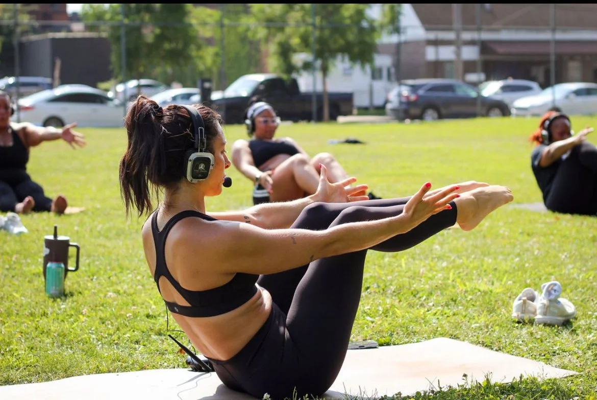 Women practicing yoga outdoors on a sunny day, sitting on yoga mats on the grass, with cars and trees in the background.