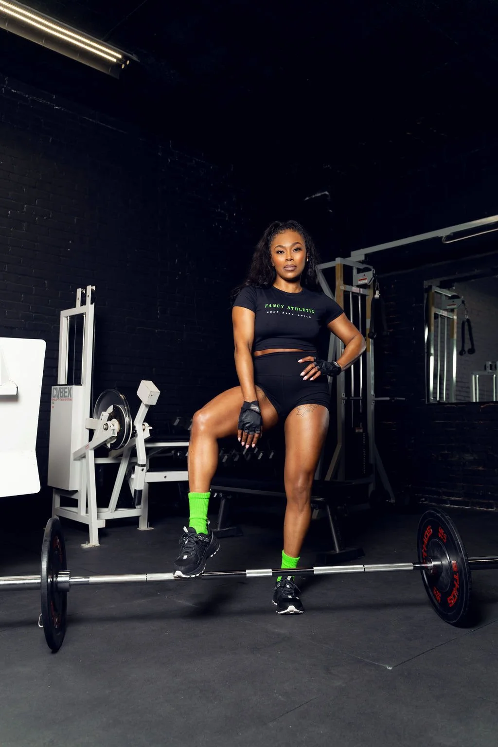 A woman in workout attire posing in a gym, standing on one leg with her foot on a barbell, black brick wall background, fitness equipment around her.