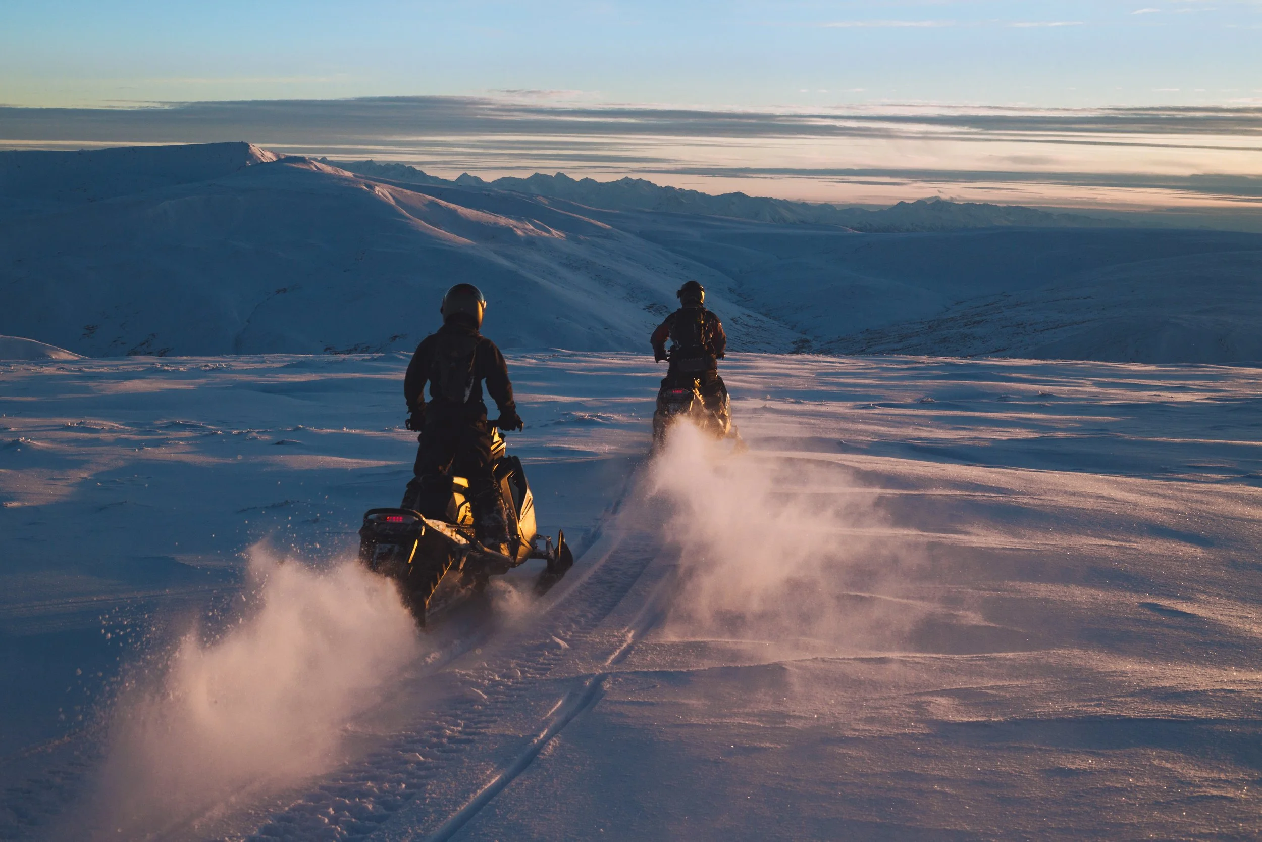 Two people riding snowmobiles across snow-covered landscape during sunset or sunrise with mountains and sky in the background.