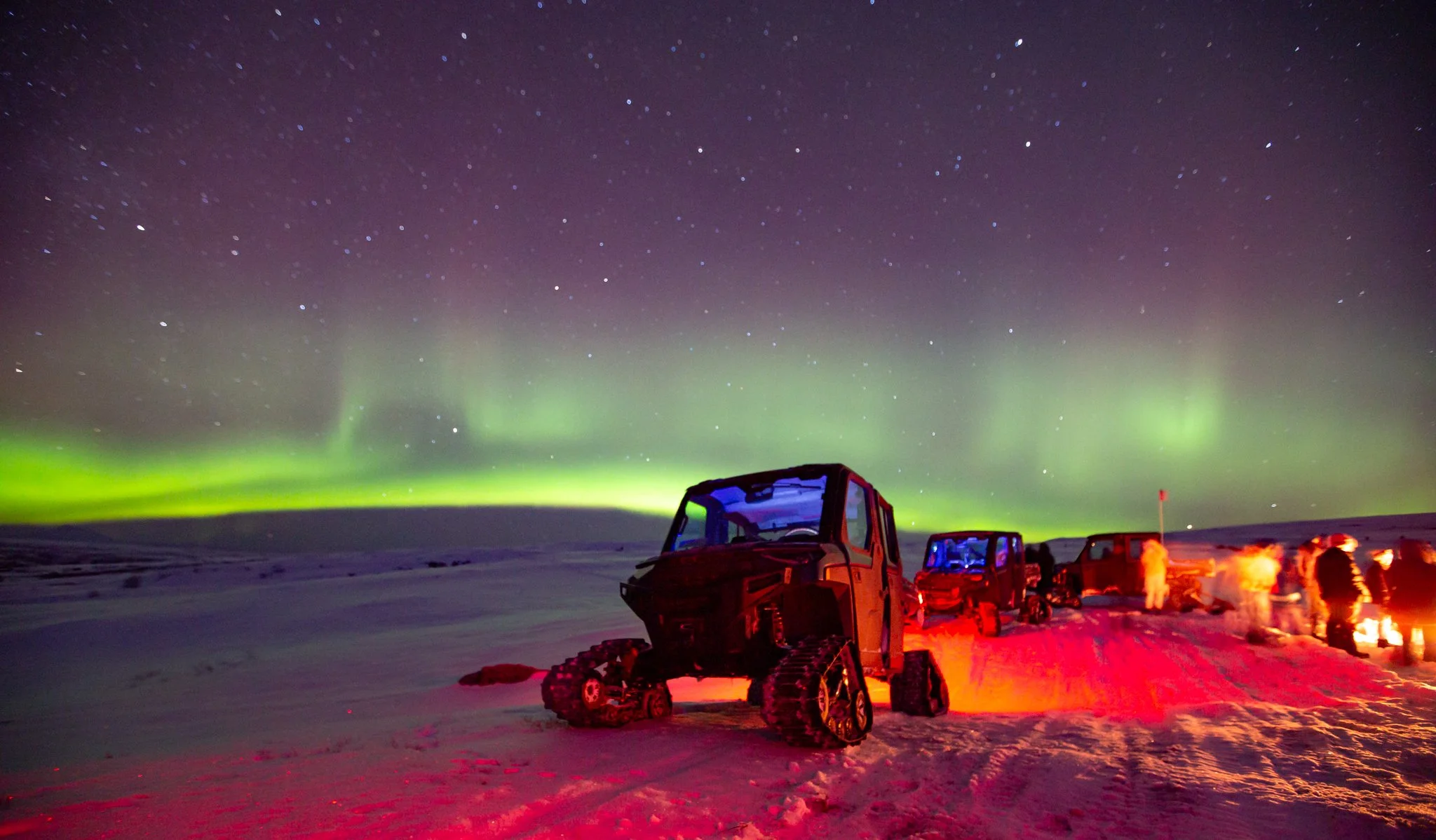 Snowy landscape at night with the Northern Lights in the sky, a small tracked vehicle in foreground, and several people around a campfire in the background.