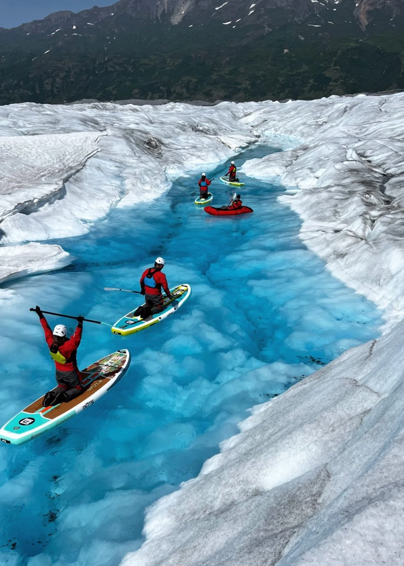 Group of people in red and blue gear on paddleboards and an inflatable raft navigating through a glacier's icy blue meltwater.