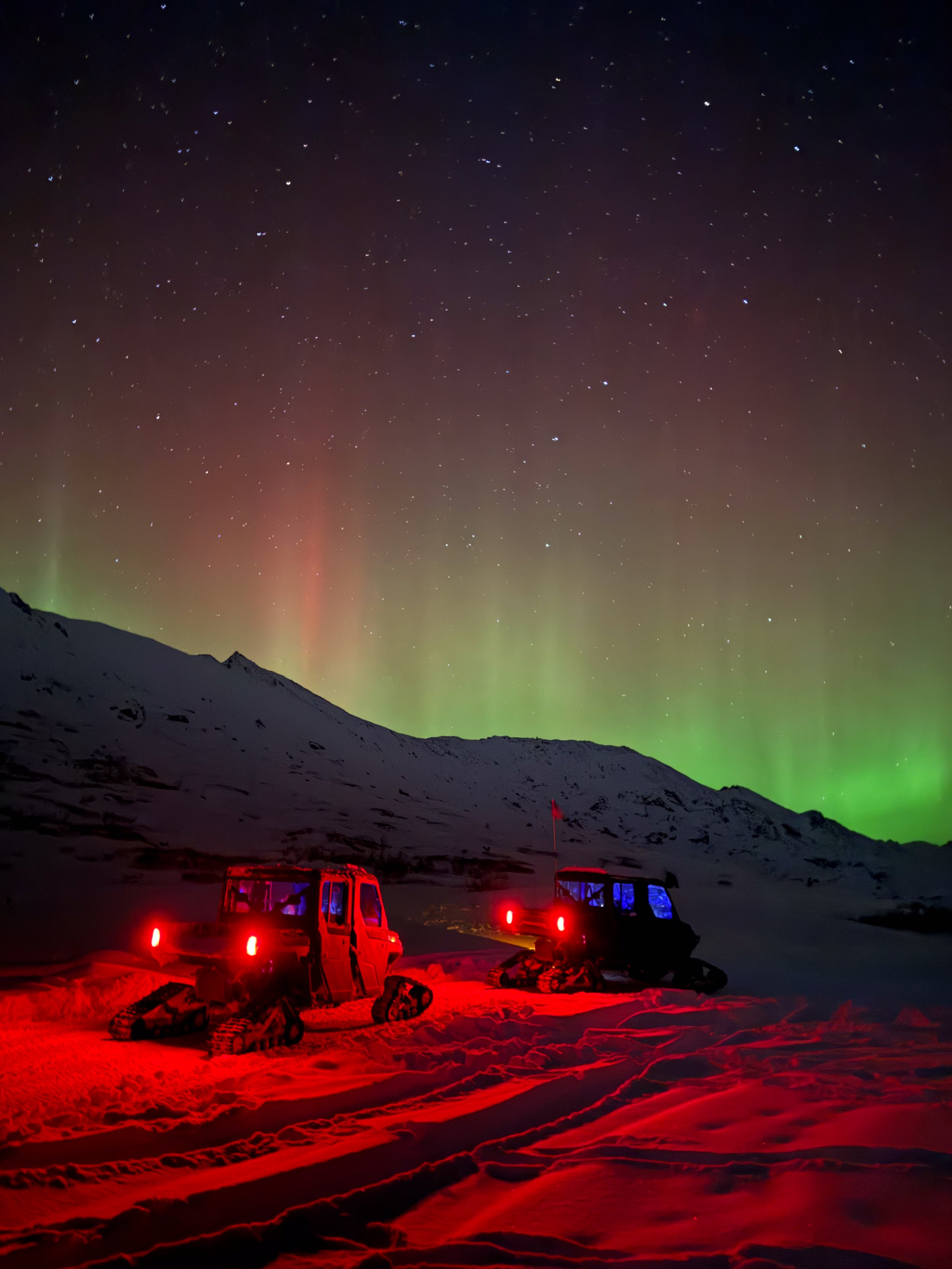Snow-covered landscape at night with snowmobiles illuminated in red lights, set against a mountain backdrop and a sky displaying the Northern Lights, stars, and celestial phenomena.