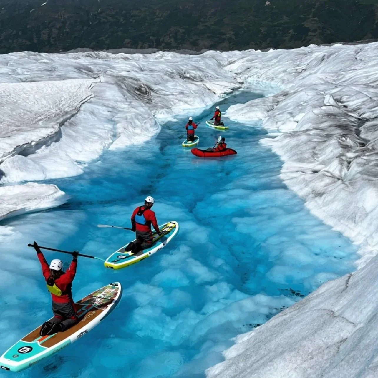 Group of five people in red and black gear riding on paddleboards and inflatable boats on a narrow, icy blue glacial river surrounded by ice and snow.