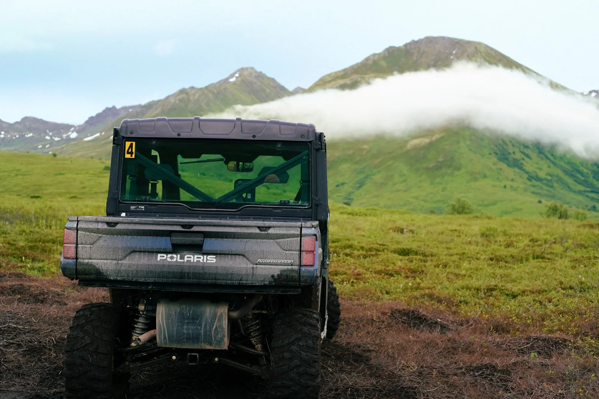 Polaris off-road vehicle parked on grassy terrain with mountains and low clouds in the background.