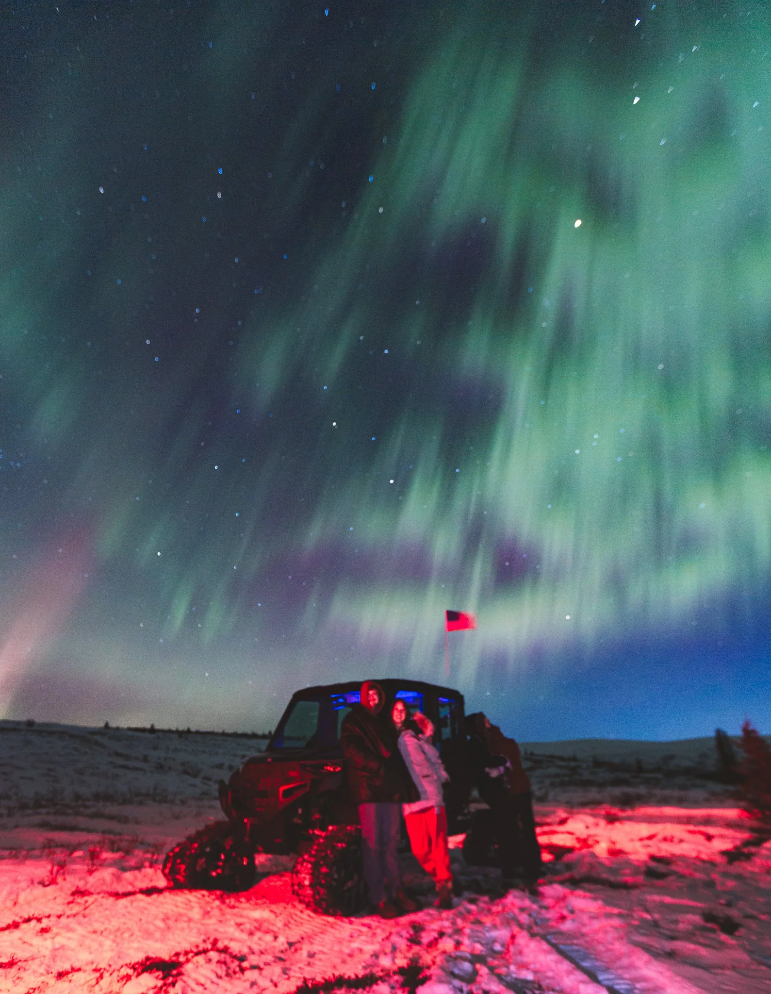People standing near a vehicle in snowy landscape at night under the Northern Lights with a visible red flag.