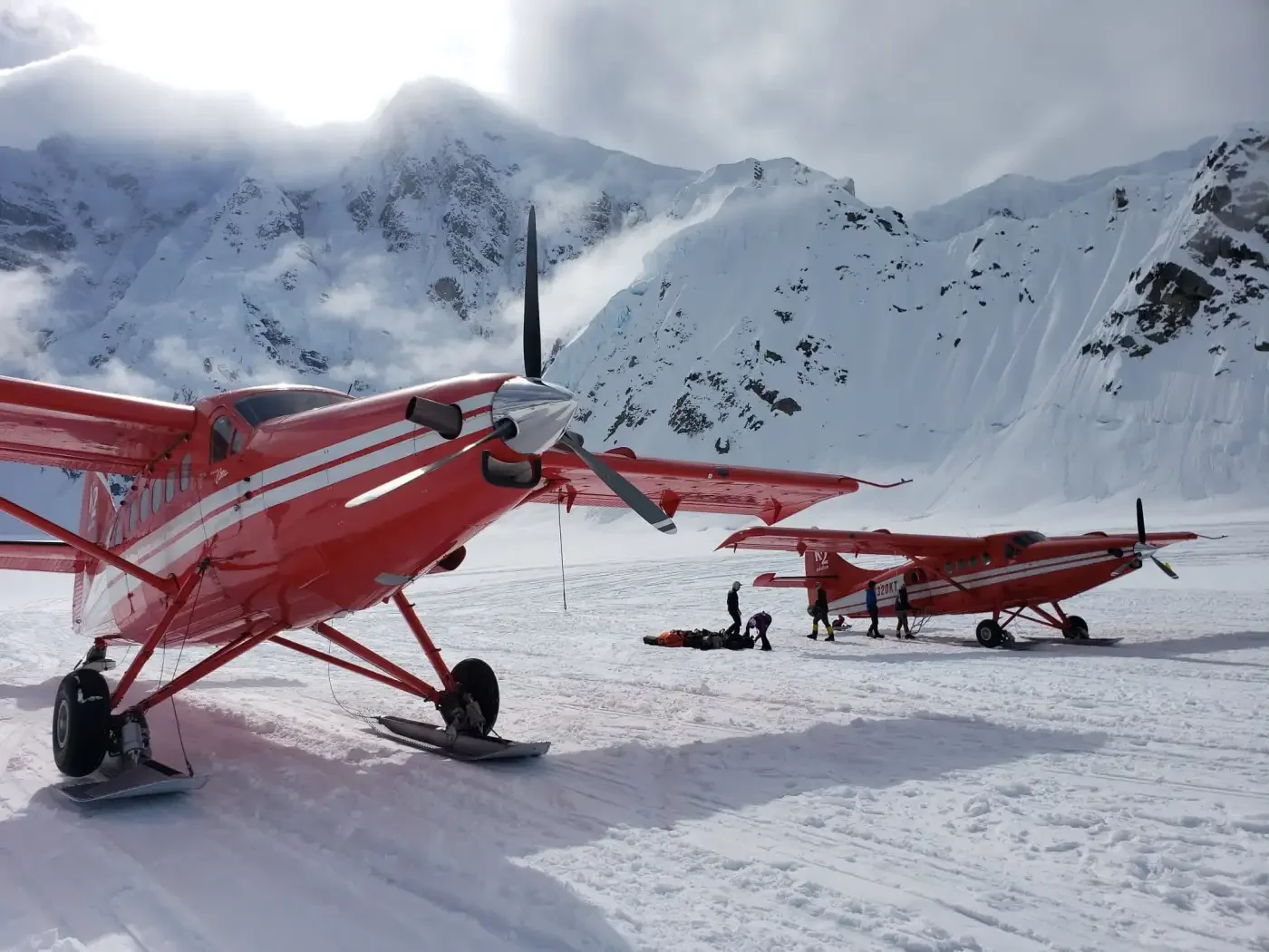 Two orange ski planes on snow with mountains in the background, and people attending to luggage or equipment near the planes.