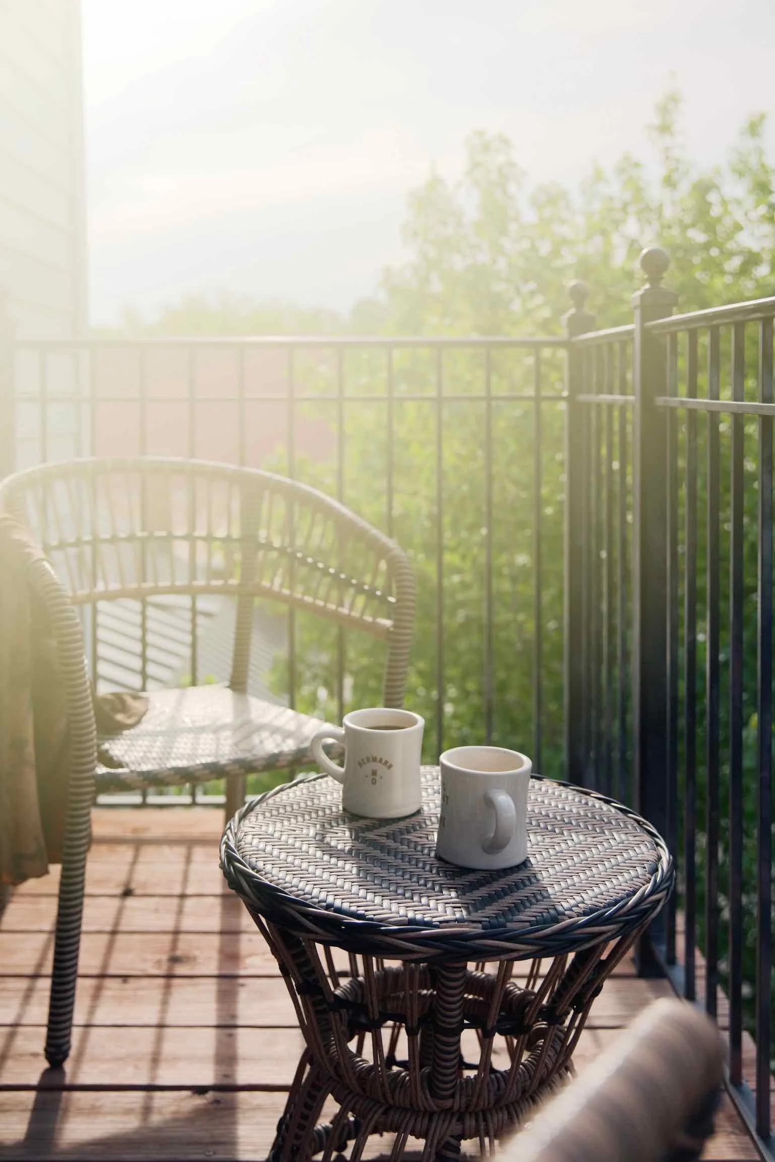 Morning on a balcony with two white coffee mugs on a wicker table, a wicker chair, and greenery in the background.