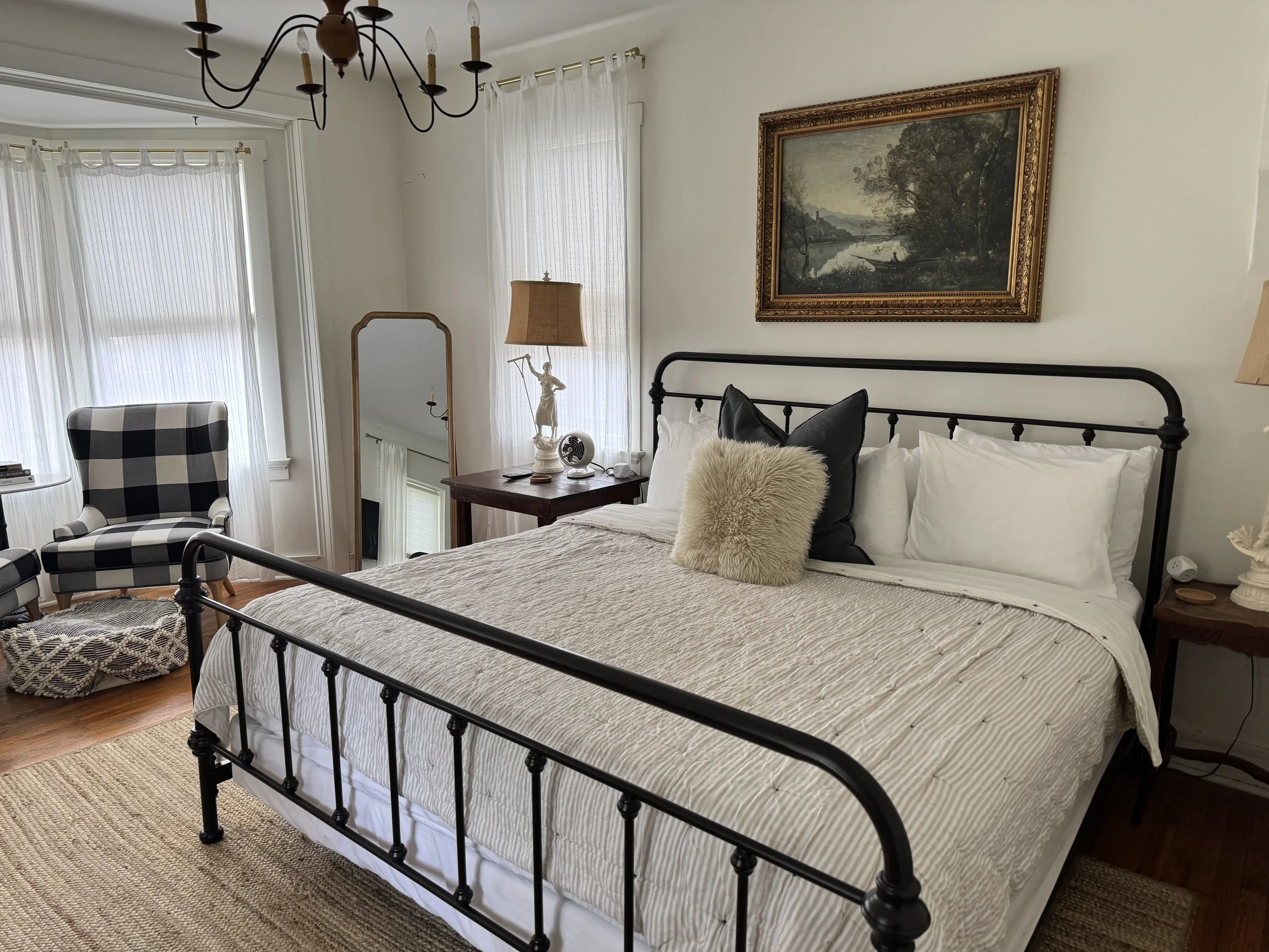 A bedroom with a black metal bed frame, white bedding, and decorative pillows, a wooden nightstand with a lamp and statue, a checkered armchair, a standing mirror, and a framed landscape painting above the bed.