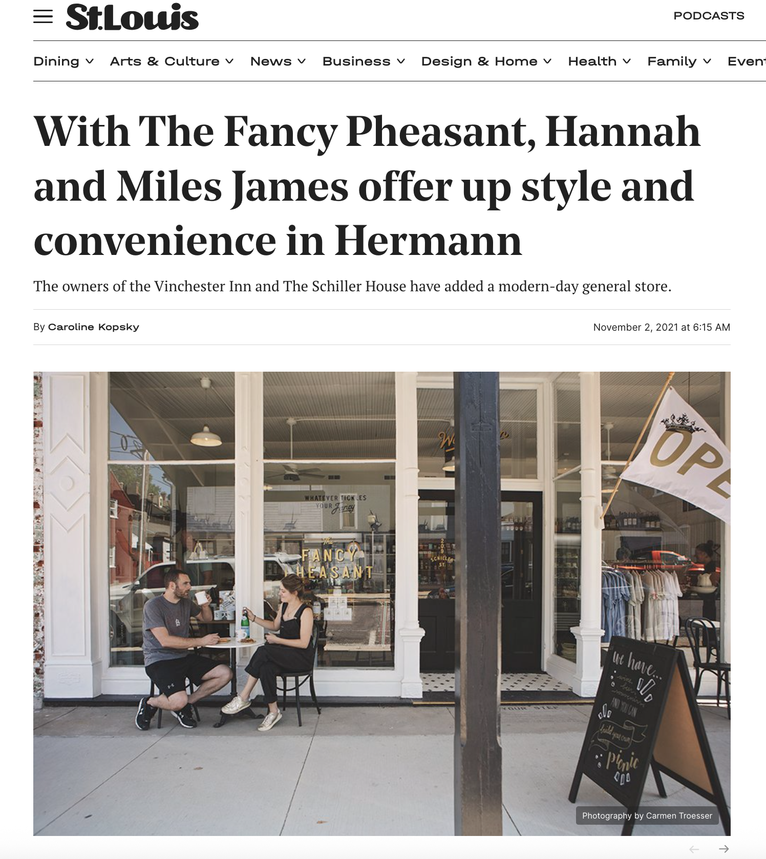 Two beds with white bedding and yellow pillows, separated by a wooden nightstand with a fan and note, against a dark wall with a framed black and white photograph and wall-mounted lamps on each side.