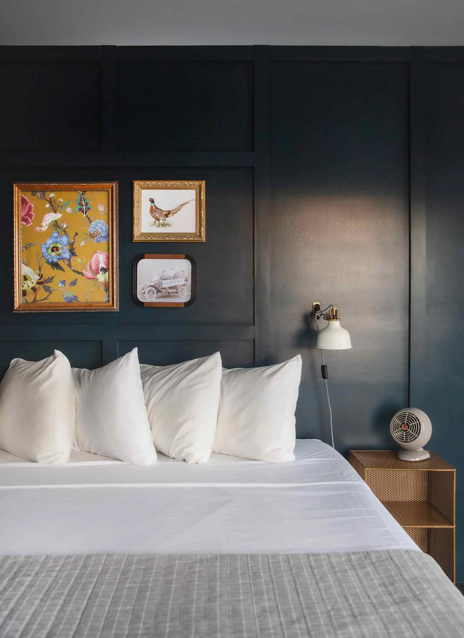 A modern bedroom with a dark blue accent wall decorated with framed artwork, including floral, bird, and vintage car images. A bed with white pillows and a gray bedspread is visible, with a white bedside lamp and a small fan on a wooden nightstand.
