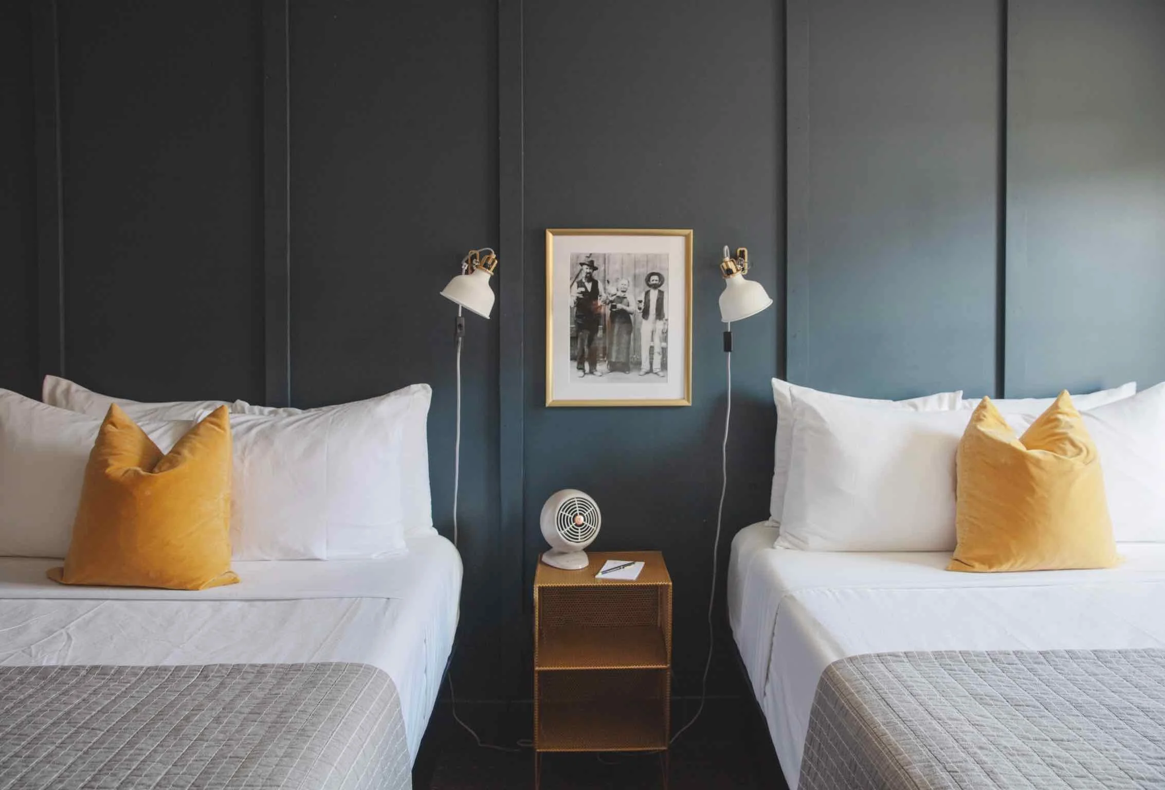 Two beds with white bedding and yellow pillows, separated by a wooden nightstand with a fan and note, against a dark wall with a framed black and white photograph and wall-mounted lamps on each side.