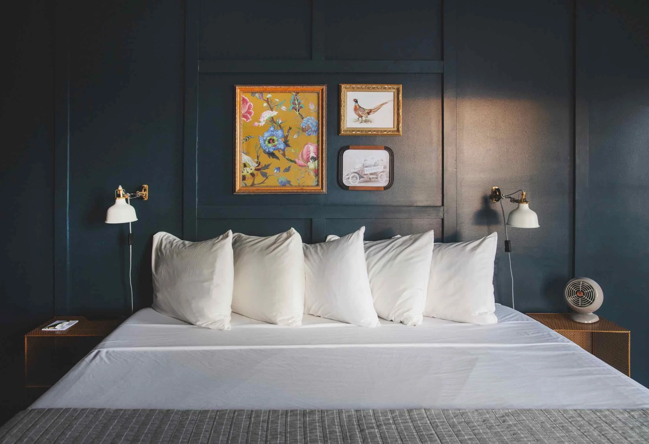 A neatly made bed with six white pillows against a dark blue wall, two wall-mounted lamps with white shades, framed artwork including a floral pattern, a bird, and a vintage car, and two small wooden side tables, one with a telephone and the other with a small fan.