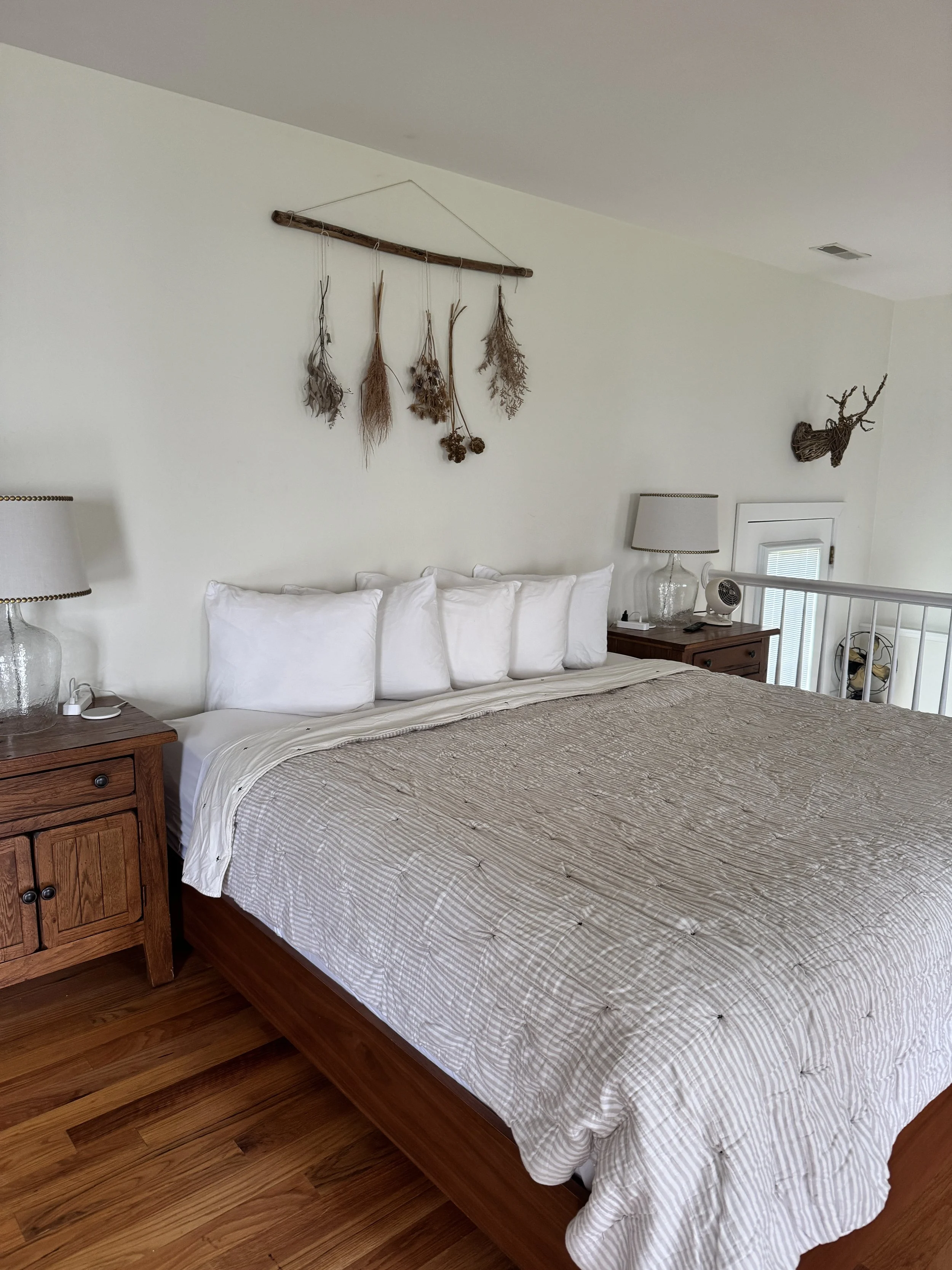 A neatly made bed with white pillows and beige bedspread in a rustic bedroom. Two wooden nightstands with glass lamps, a small fan, and decorative wall hangings of dried herbs and a mounted wooden deer head are also visible.