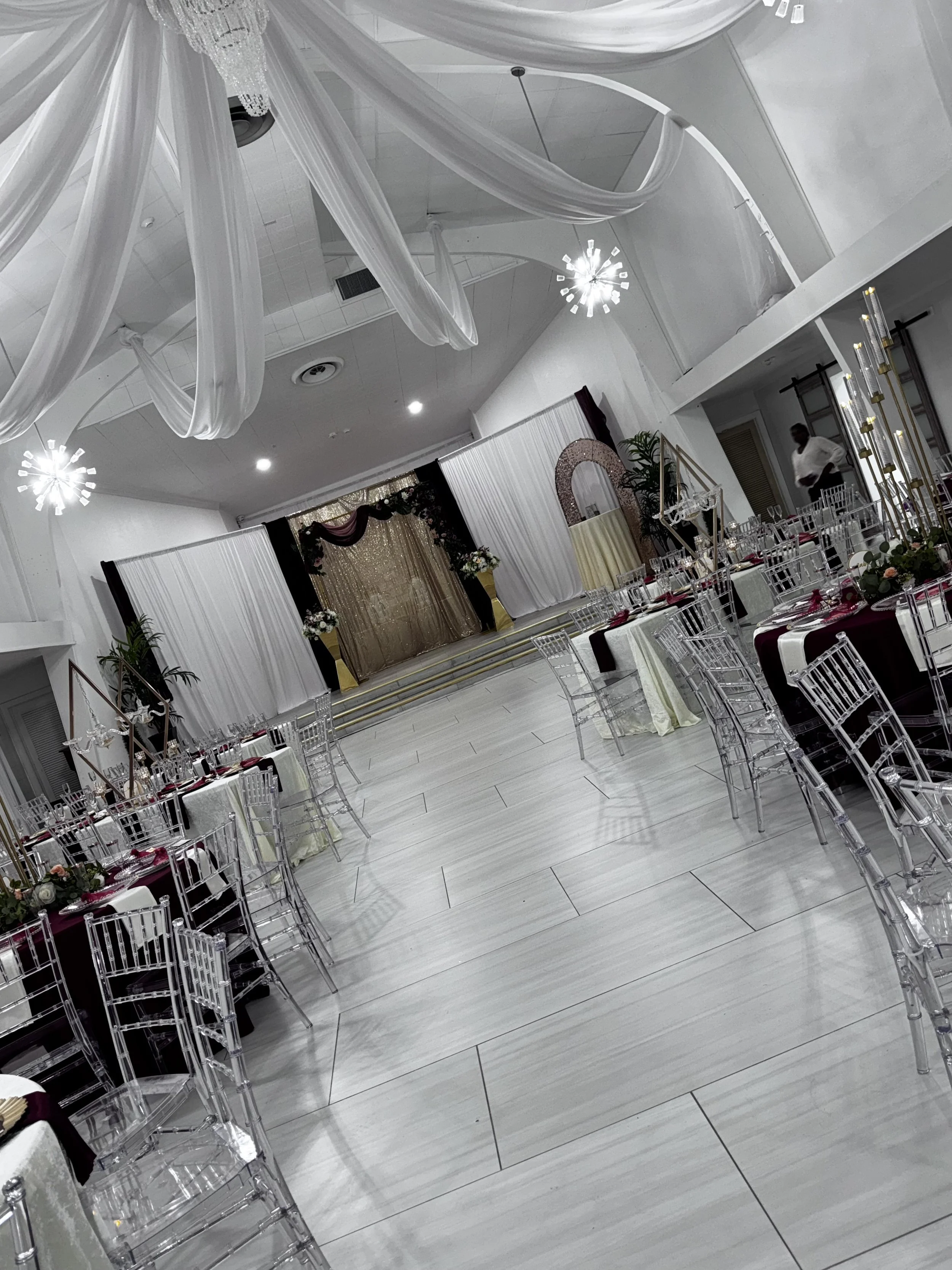Elegant event hall decorated with white drapery, chandeliers, tables with maroon and white table settings, clear chairs, and a stage backdrop with curtains and floral arrangements.