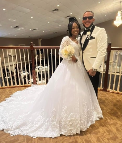 A bride and groom smiling on their wedding day, with the bride in a white wedding dress holding a bouquet and the groom in a white tuxedo with black accents, standing indoors near a wooden railing.