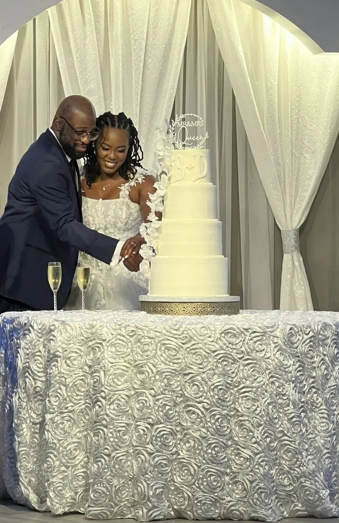 A bride and groom in wedding attire cutting a white wedding cake together at their celebration, with champagne glasses on the table and elegant backdrop decorations.