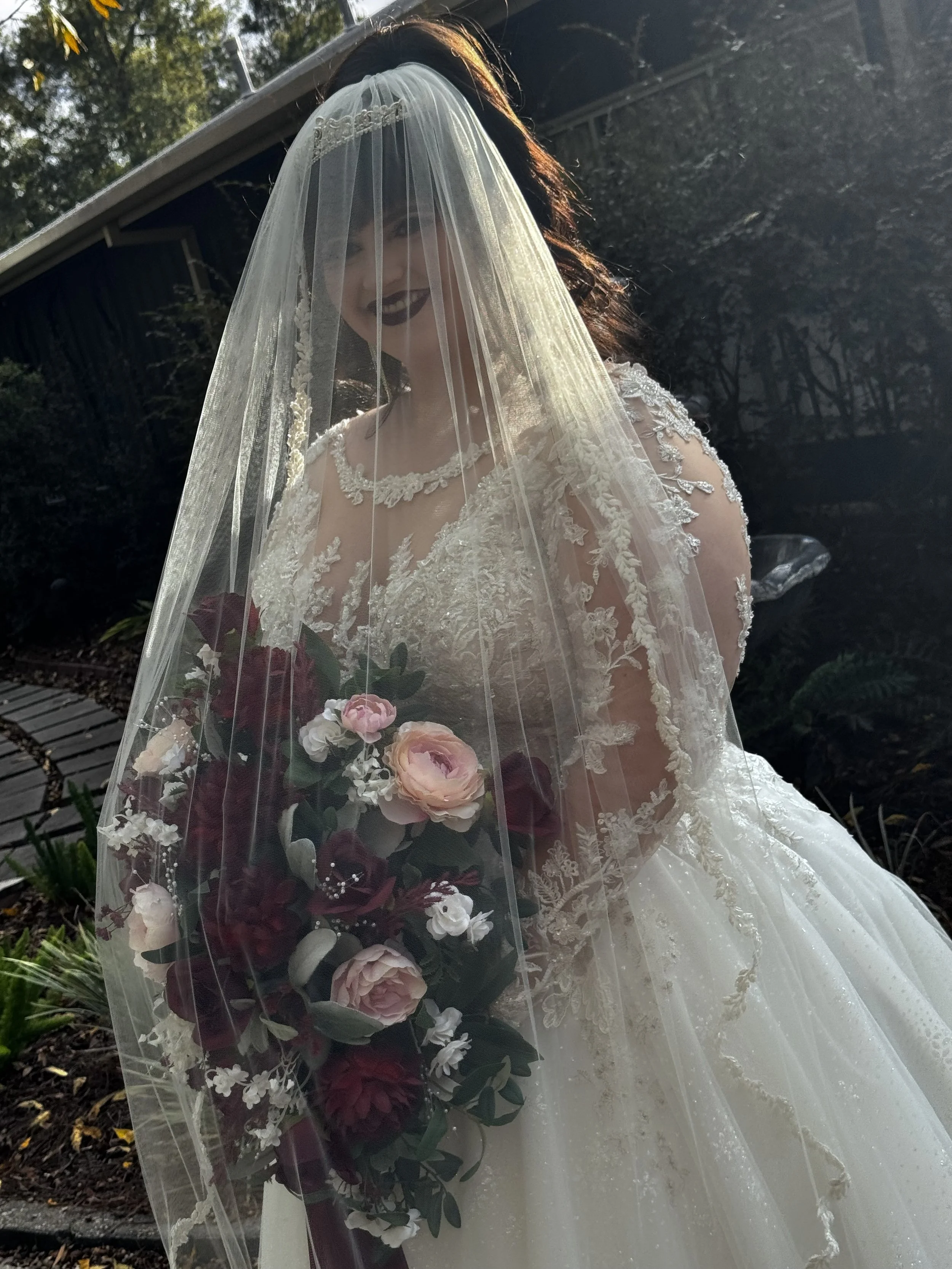 A bride in a wedding dress with lace details, holding a bouquet of pink, white, and burgundy flowers, smiling under a veil in an outdoor garden setting.