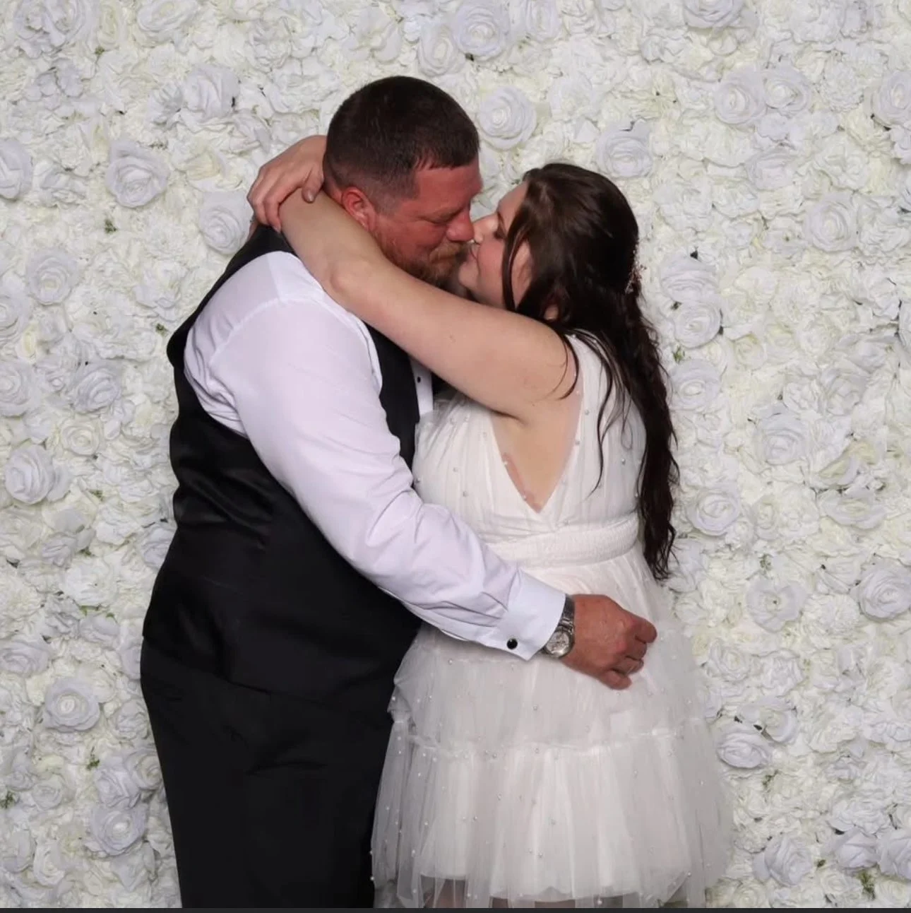 A couple in wedding attire kissing and embracing each other against a backdrop of white roses.