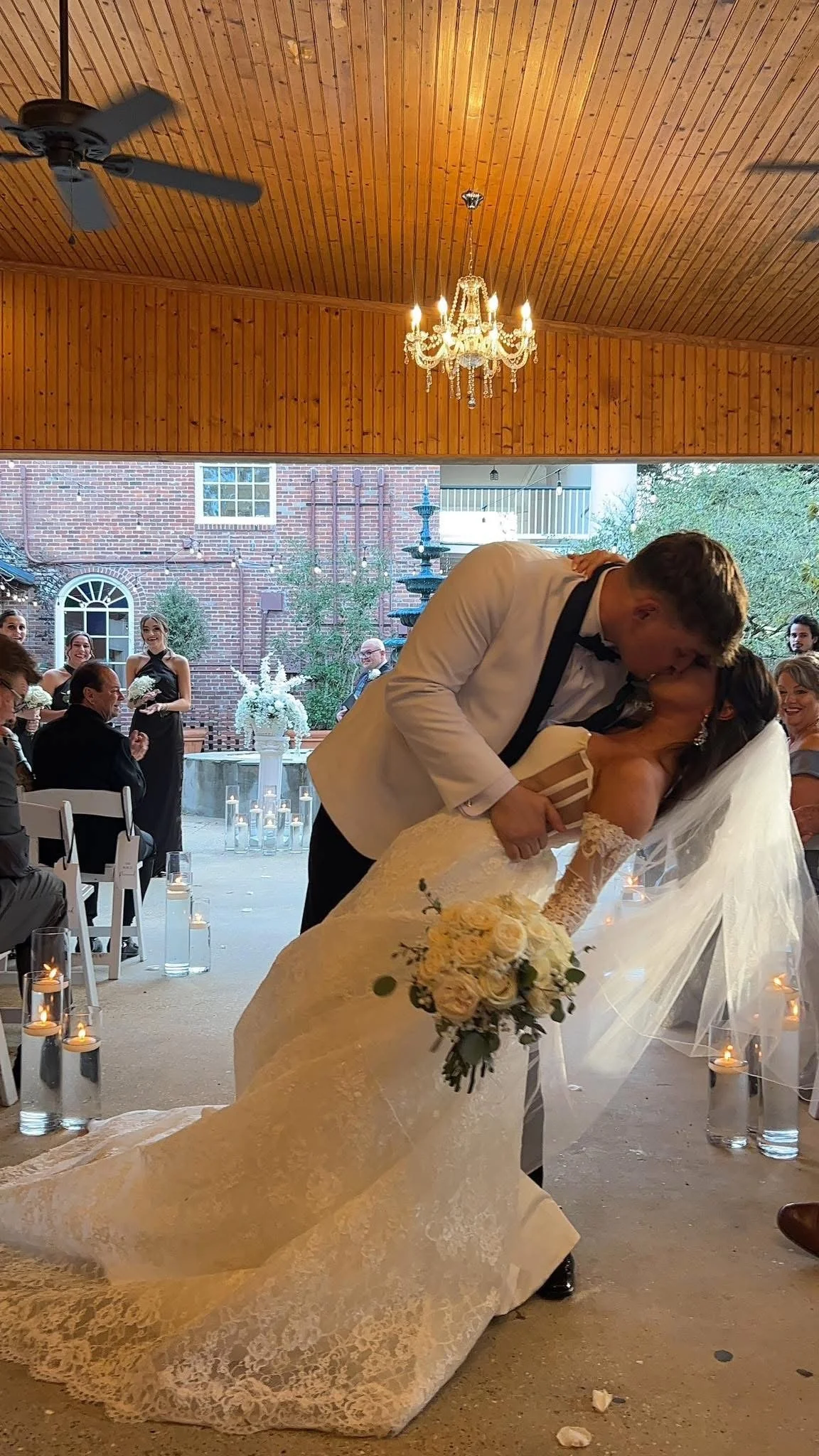 A newlywed couple sharing a kiss during their wedding reception. The groom is in a white tuxedo, and the bride is in a lace wedding dress holding a bouquet of yellow roses. Guests are seated and standing around, with candles and floral decorations visible. The setting appears to be an indoor-outdoor space with wooden ceiling and celebration atmosphere.
