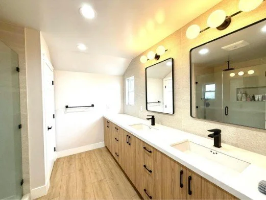 A modern bathroom with a long wooden vanity, two sinks, black faucets, and three mirrors. The space is brightly lit with round light fixtures above the mirrors, and there is a white wall with a towel rack and a window on the far wall.