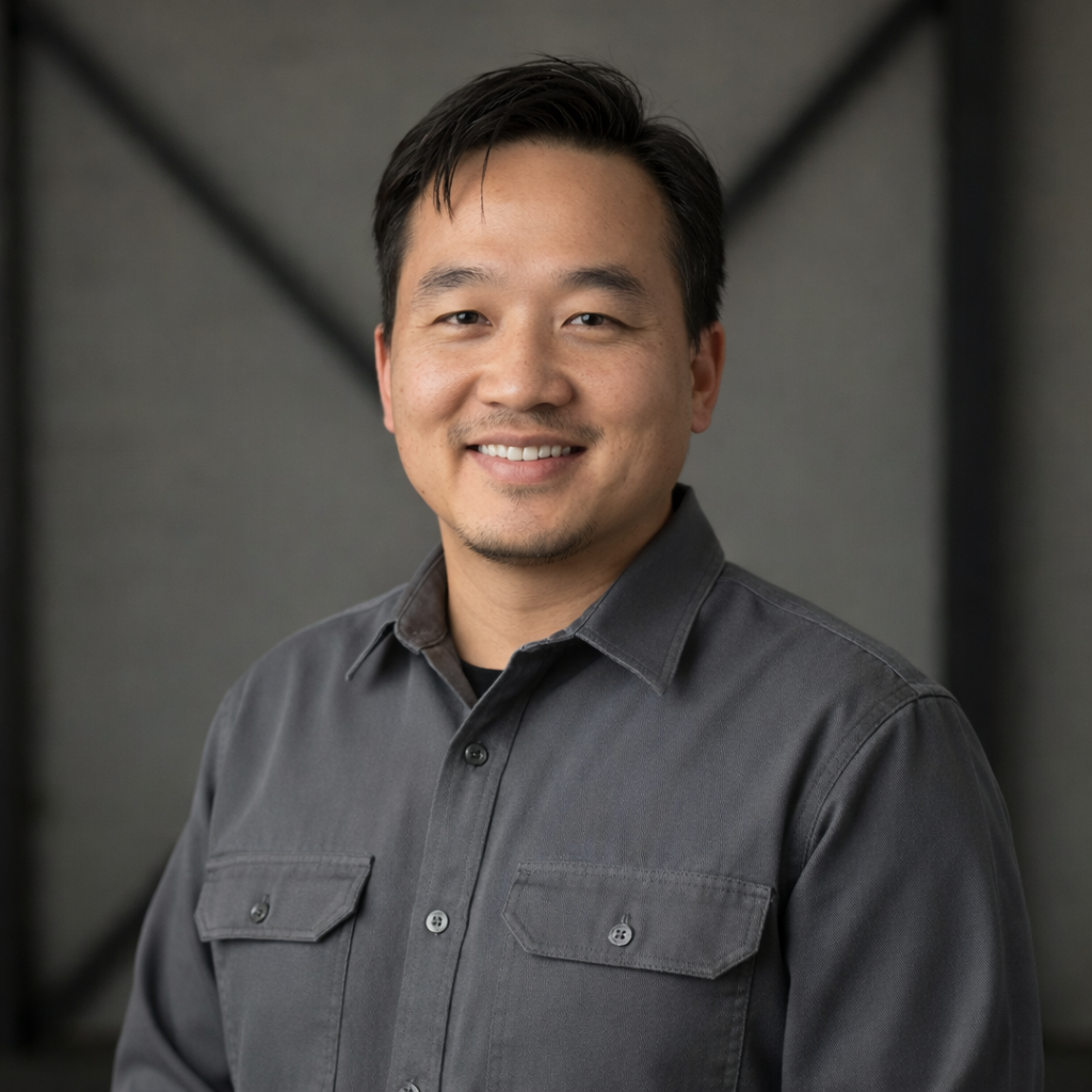 A smiling Asian man with black hair wearing a gray button-up shirt standing indoors against a blurred gray background.