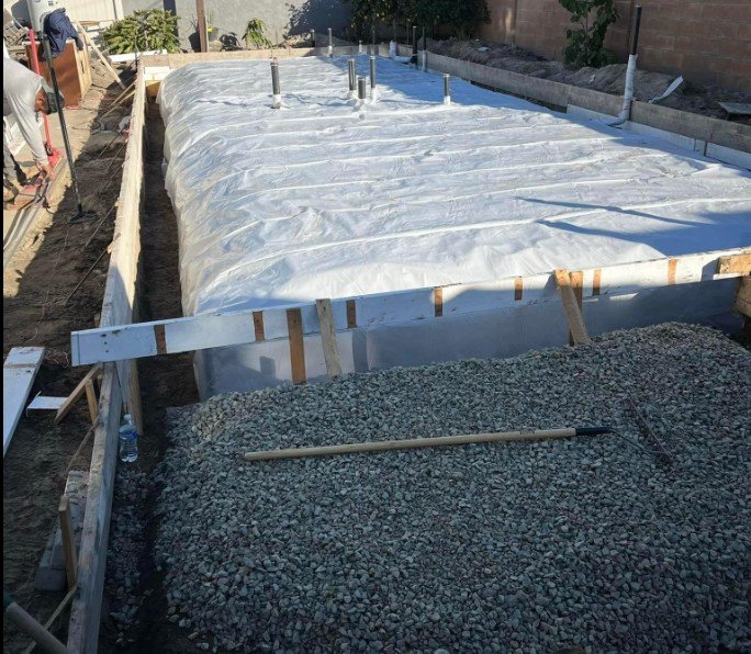 Construction site with a foundation covered in white plastic sheeting, surrounded by wooden forms and gravel, with a broom placed on top of the gravel.